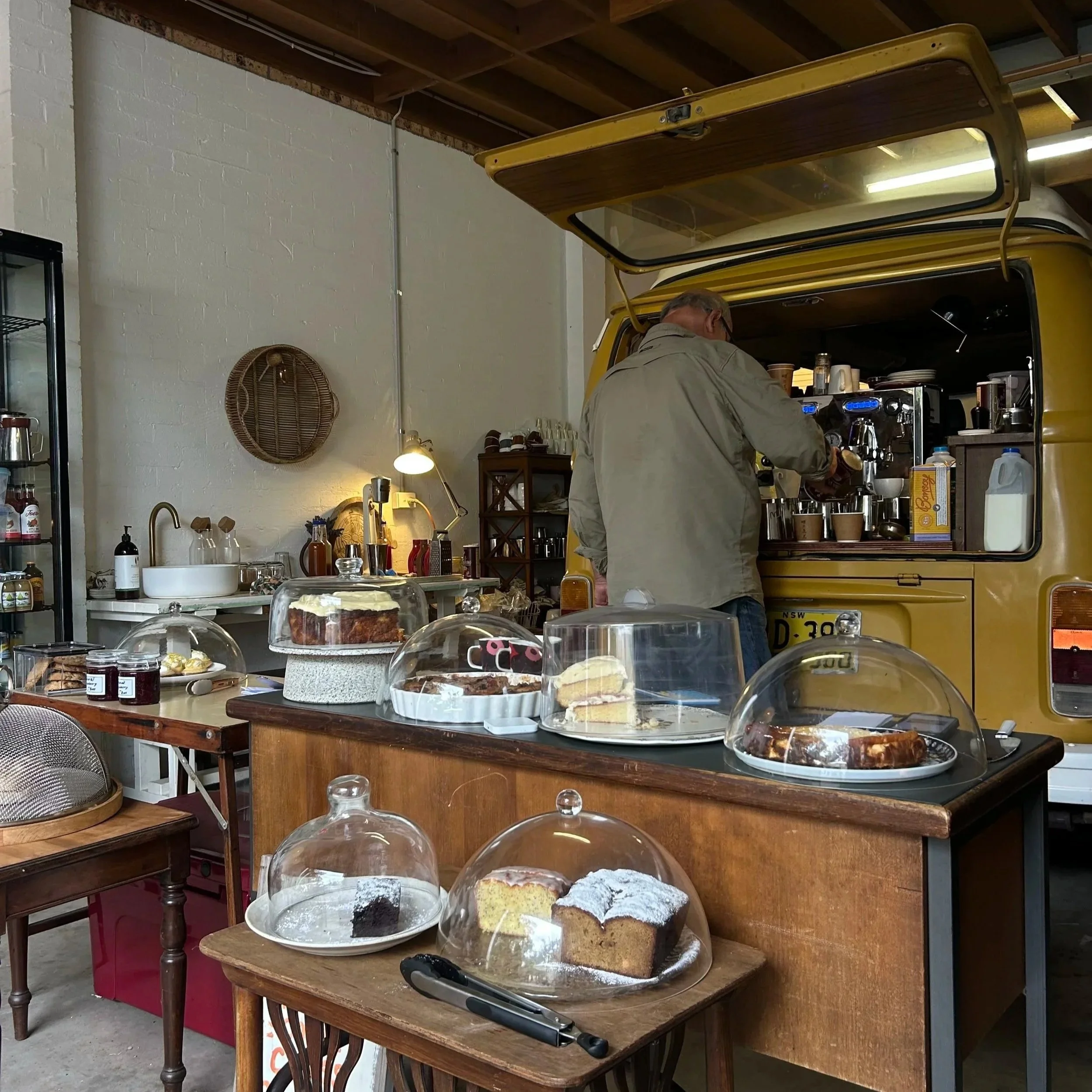 Almas Kombi Cafe with display of assorted cakes with glass covers on a wooden table, with desserts and equipment in background inside a local garage.