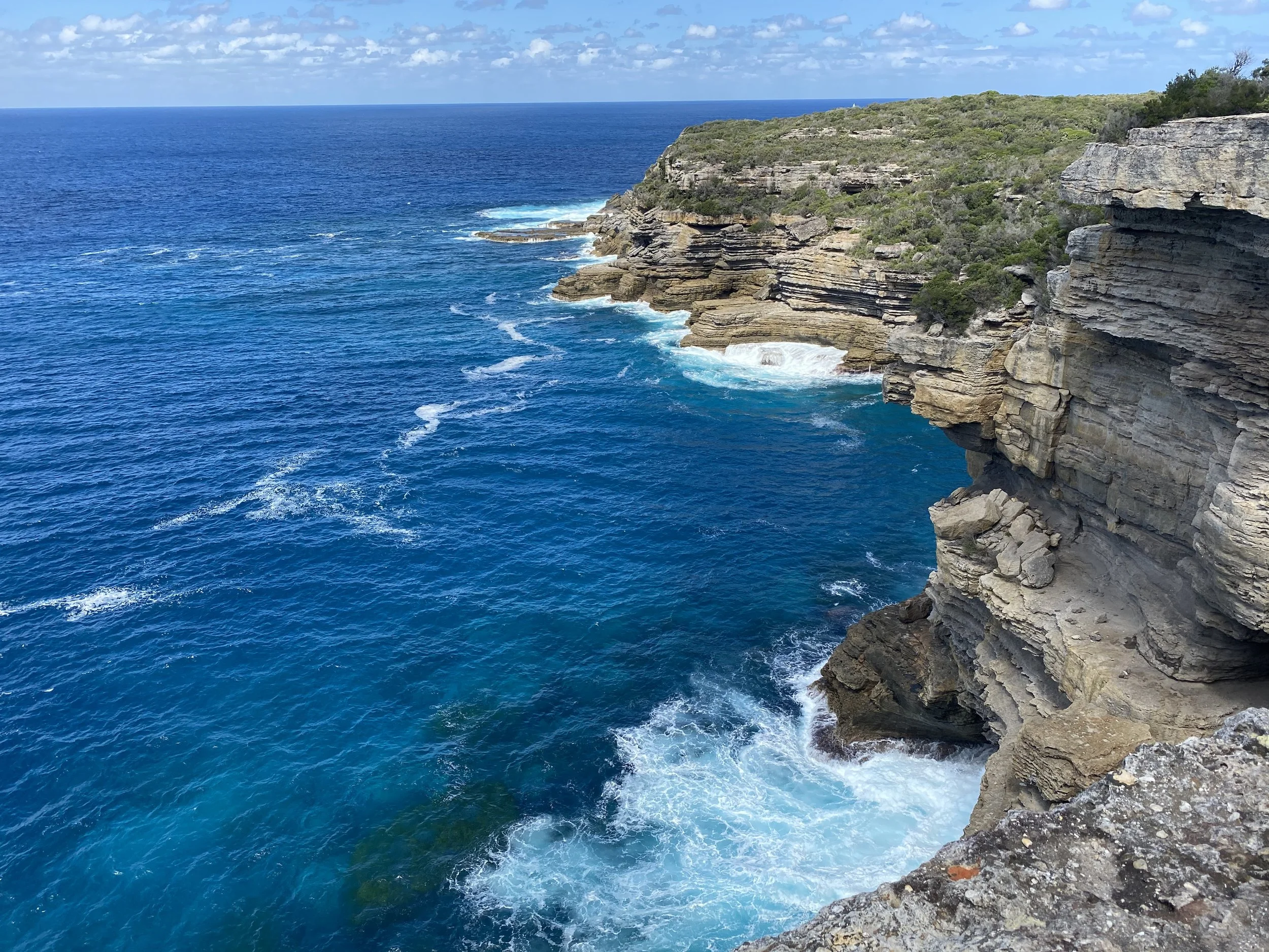Cliffside overlooking the ocean with blue water, layered rocks, and greenery under a partly cloudy sky.