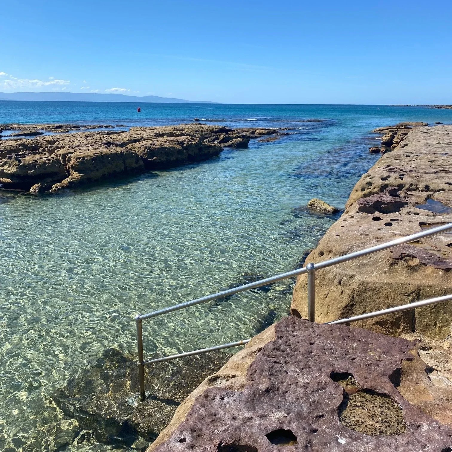 Currarong Rock Pool rocky shoreline with clear shallow water and a metal railing, extending into the ocean with blue sky with few clouds.