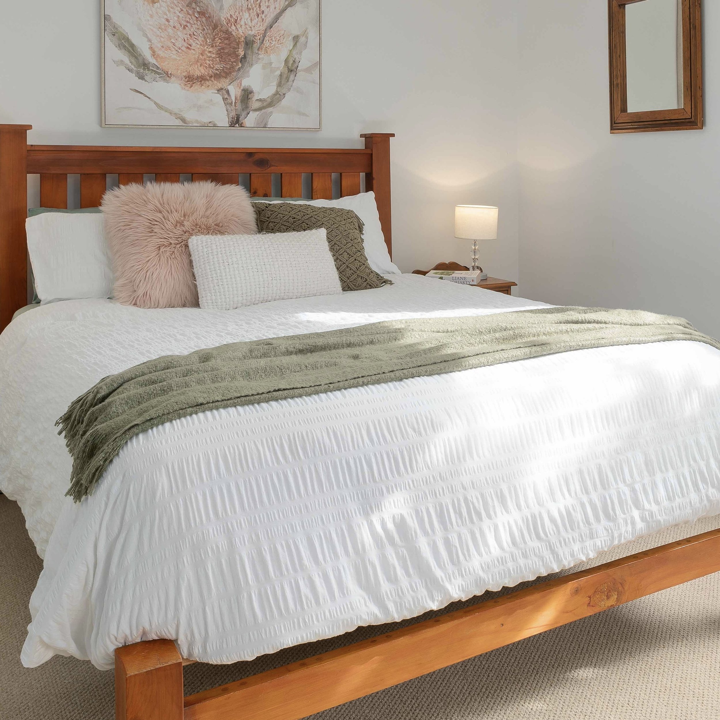 A wooden bed with a white textured bedspread, decorated with plush pillows including a pink fluffy one and a knitted beige one, with a bedside table holding a lamp and books, against a light-colored wall with framed artwork.