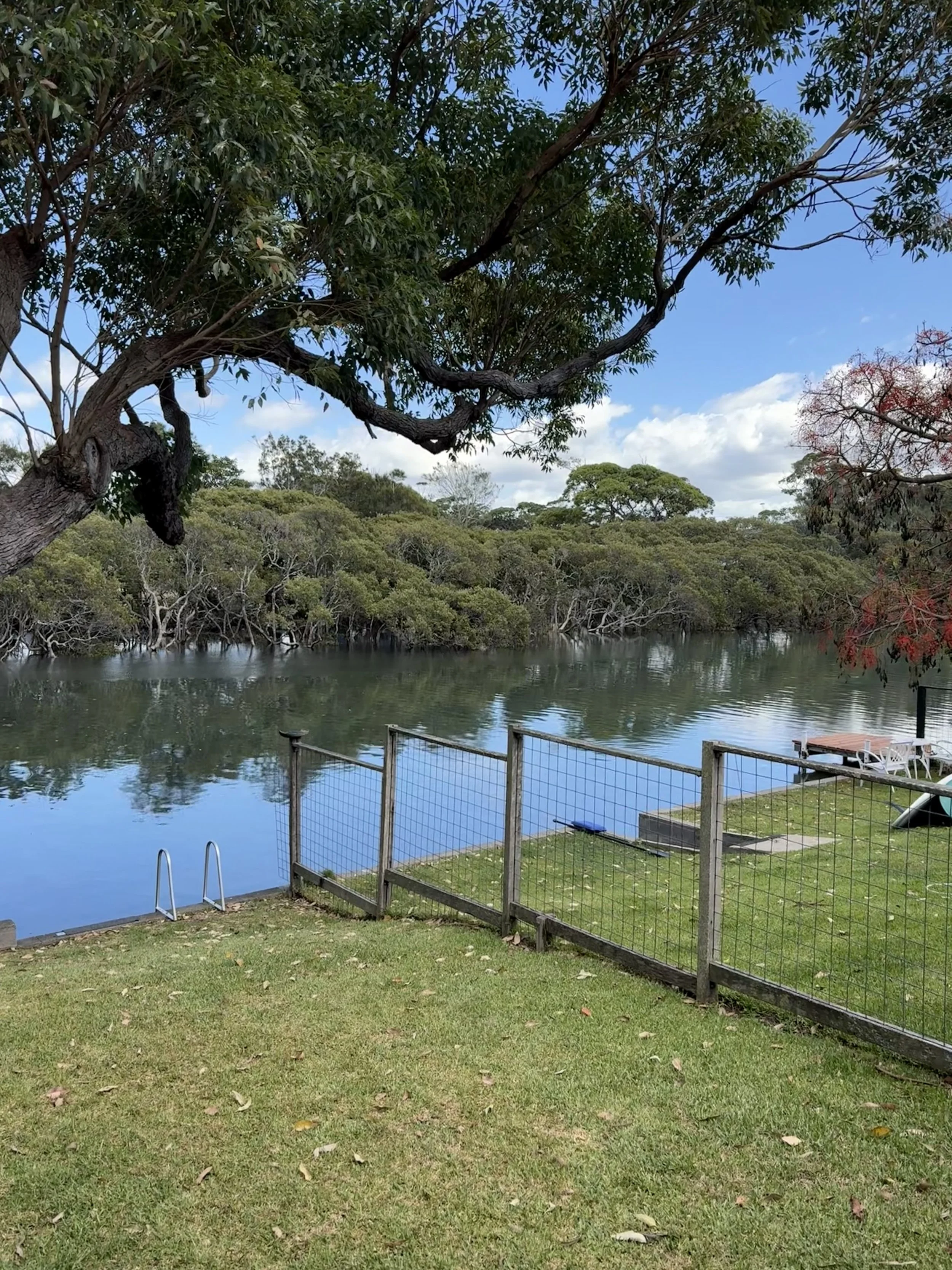 A backyard scene with a grassy area overlooking Currarong tidal creekland trees around the water. The sky is partly cloudy with a mix of blue sky and white clouds. There is a small wooden deck and outdoor furniture next door.