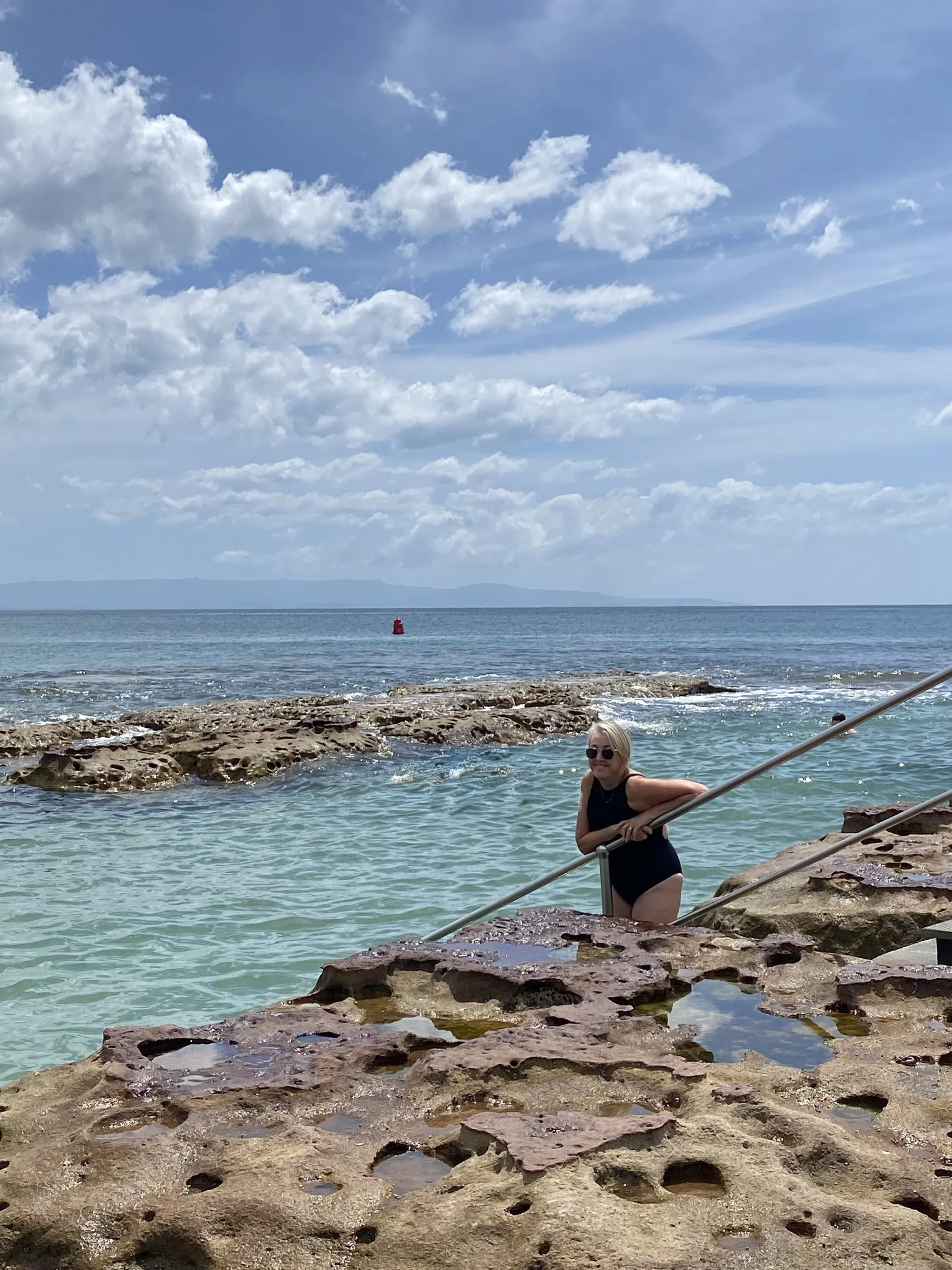 A woman in a black swimsuit with sunglasses posing on rocky steps leading into Currarong rock pool, with a red buoy floating in the water and a distant shoreline with hills under a cloudy sky.