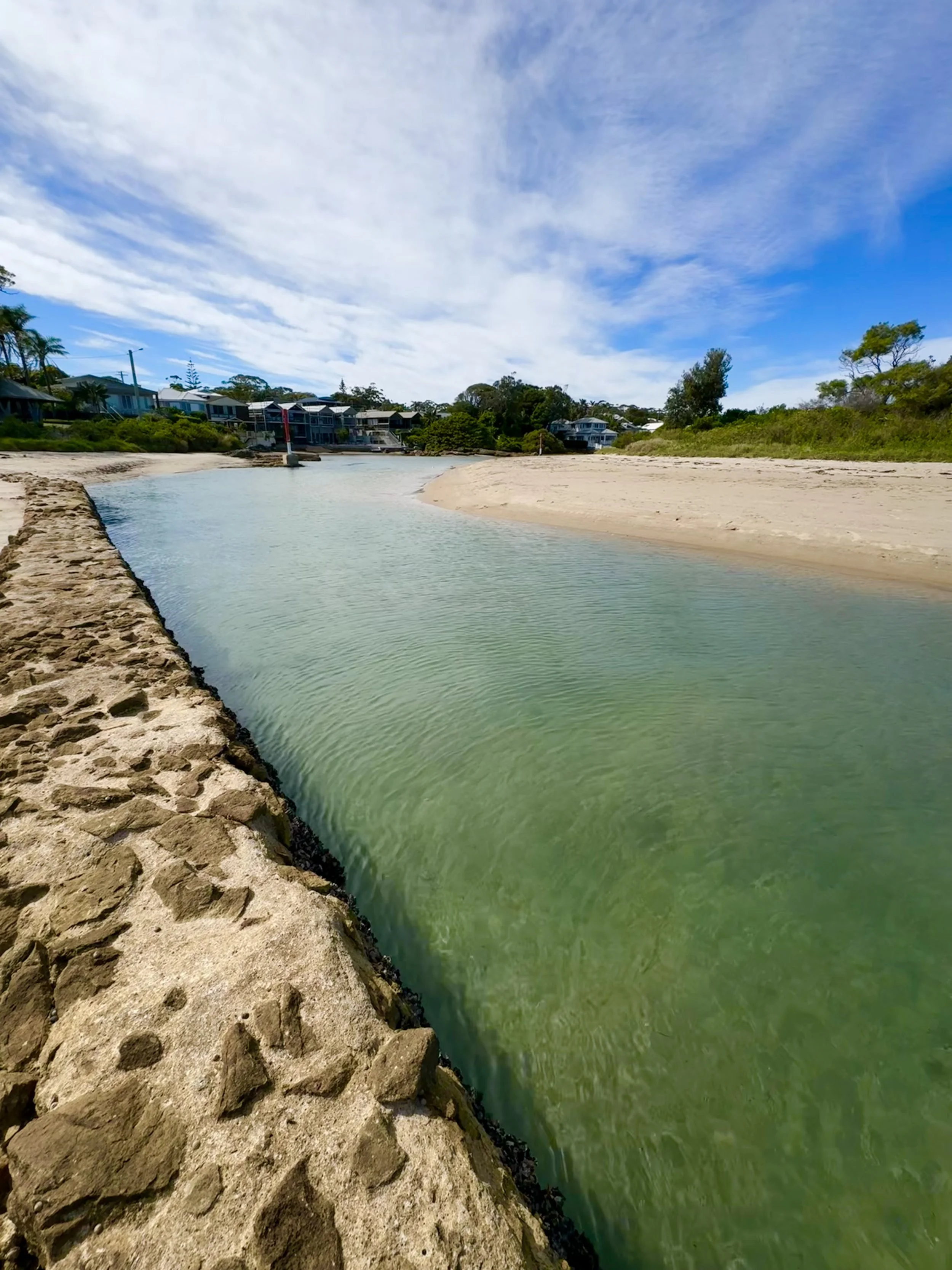 A sandy beach with a narrow water channel, houses in the background, green foliage, and a partly cloudy blue sky.