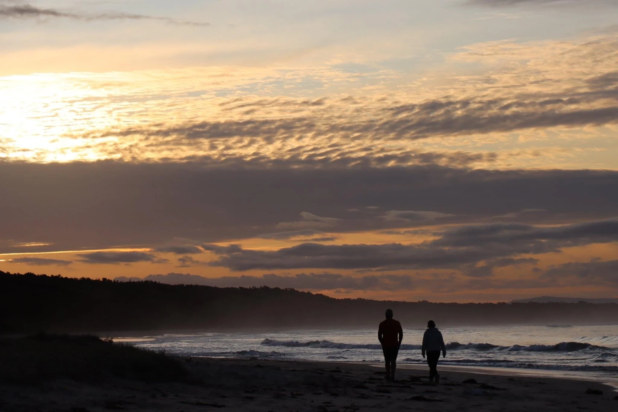 Two people walking along the shoreline at sunset, with a cloudy sky and dark silhouette of trees in the background.