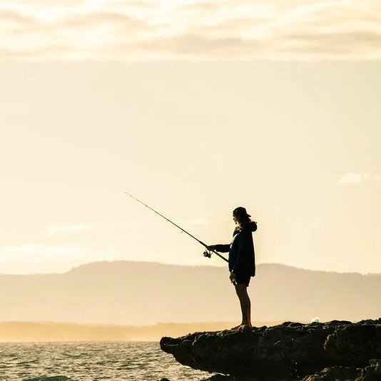 Person fishing on a rocky shoreline at sunset.