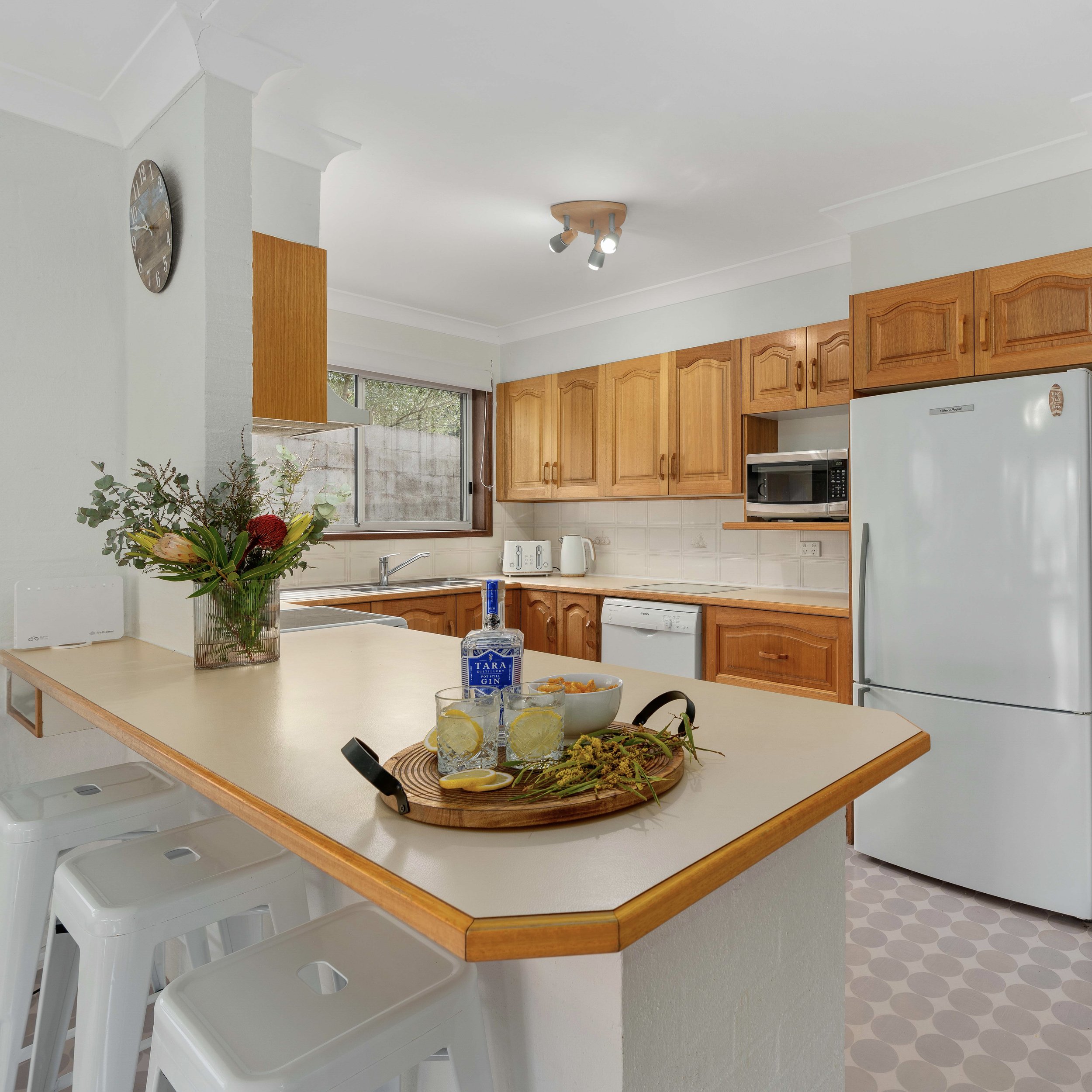 Kitchen with wooden cabinets, white appliances, a window, and a white countertop with a vase of flowers and drinks.