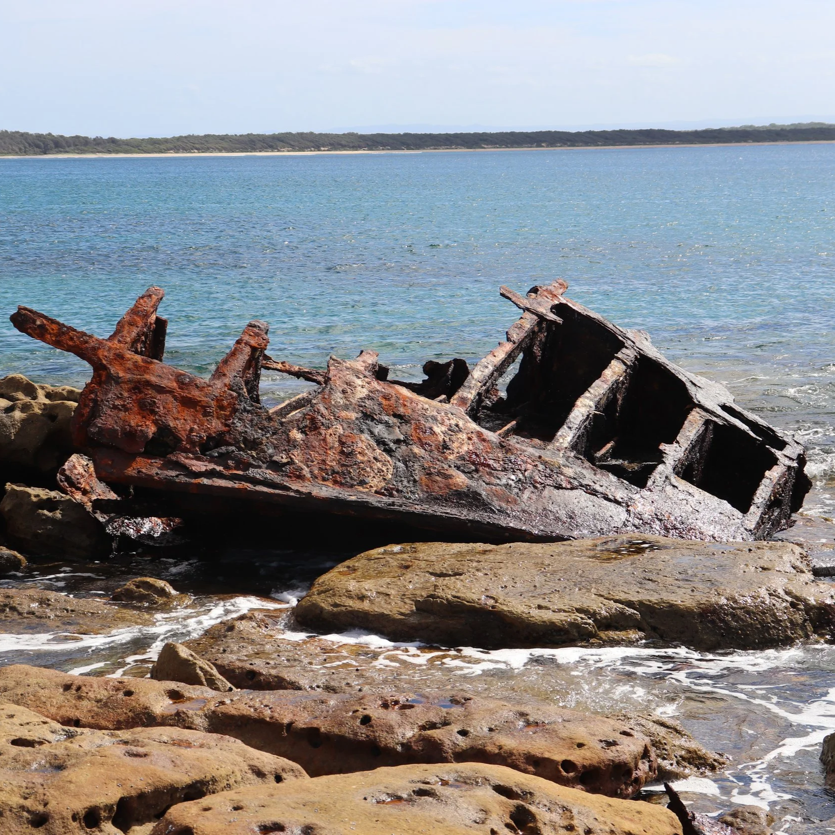 Rusty, wrecked ship remains on a rocky shoreline by calm blue water with a Shoalhaven coast in the distance.