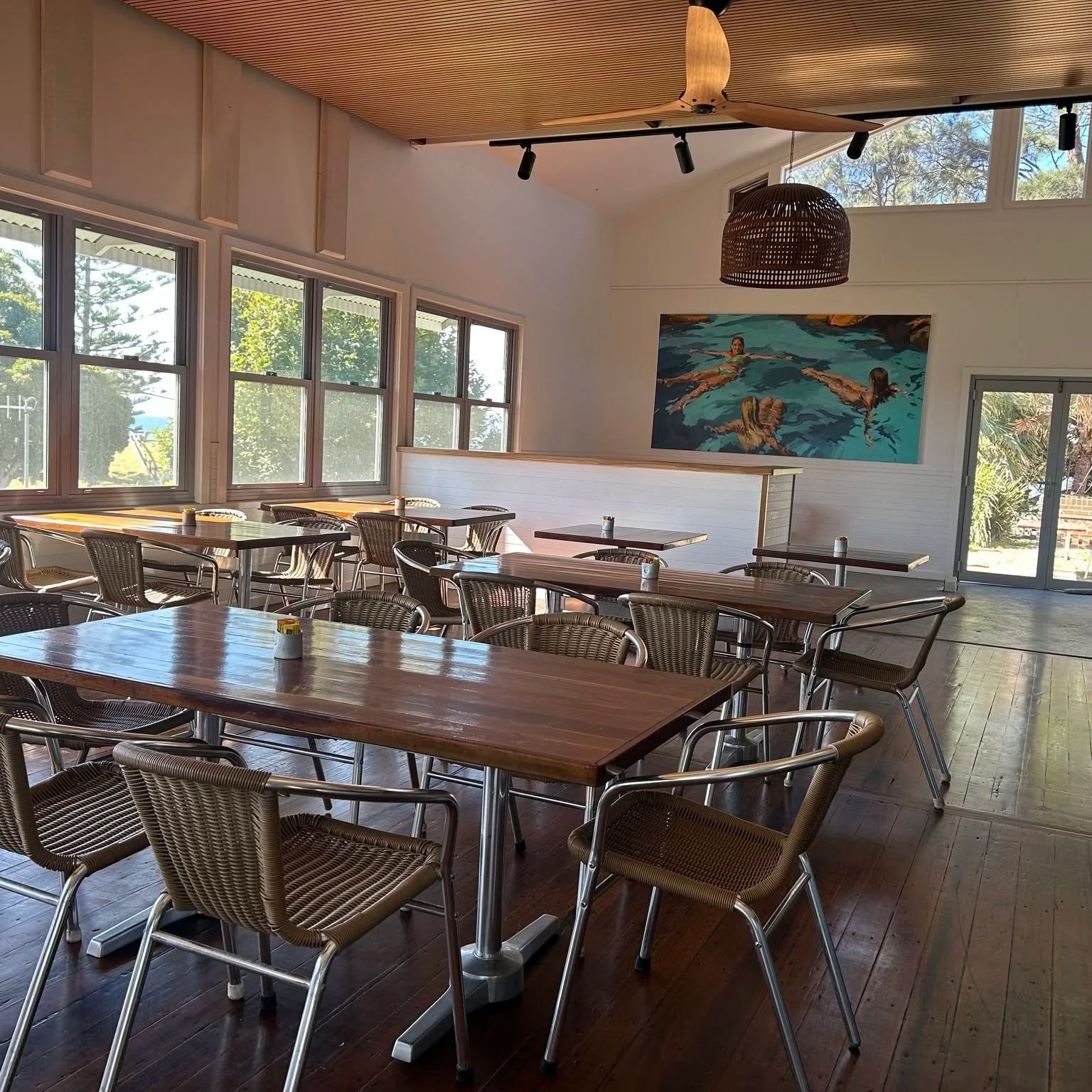 Zest at the Beach cafe with wooden tables and wicker chairs, large windows letting in natural light, a wicker pendant light, an Robyn Gibson painting of people swimming.