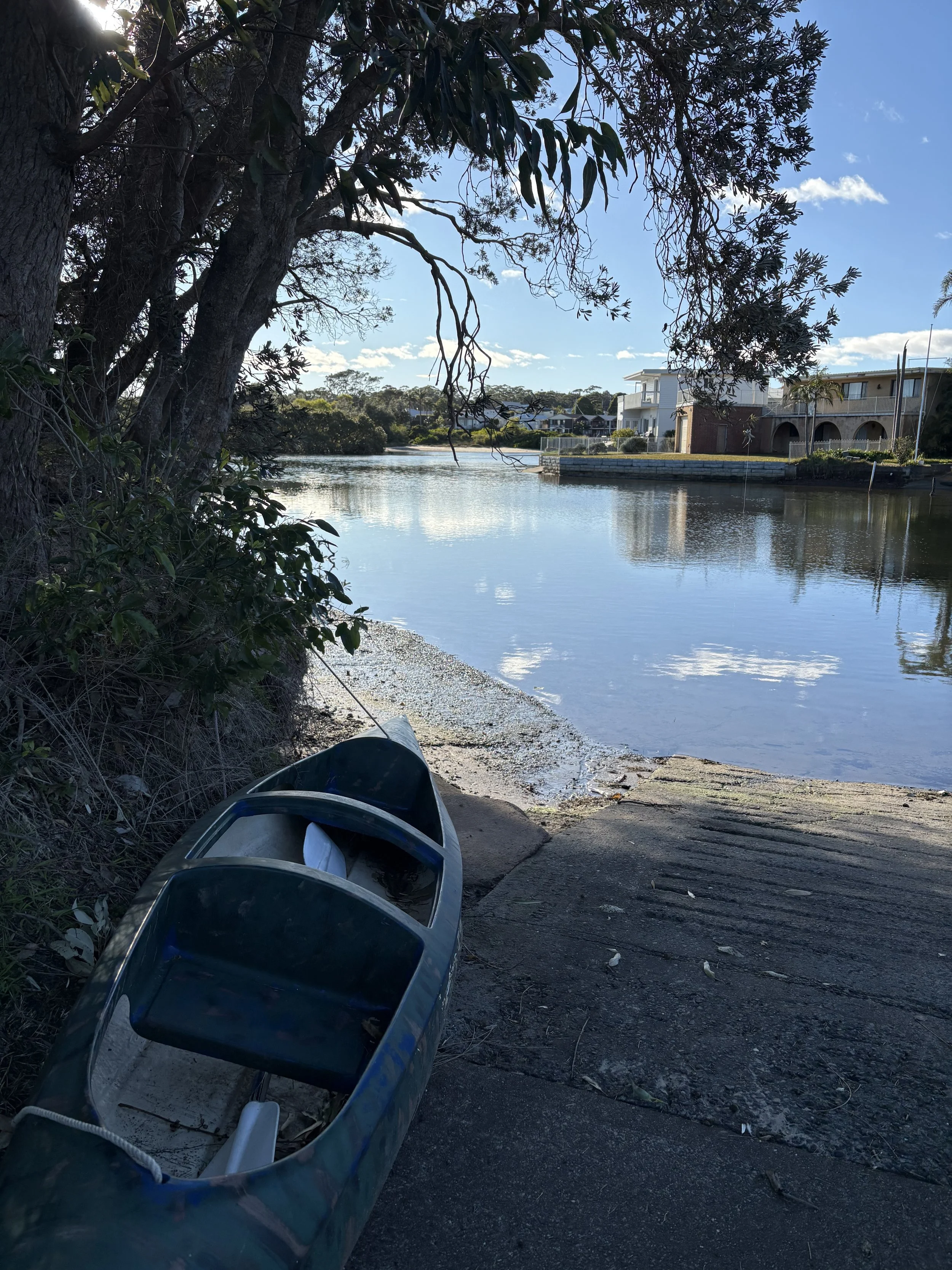 A creek scene with trees, calm water, and houses in the background. A kayak is partly visible in the foreground.