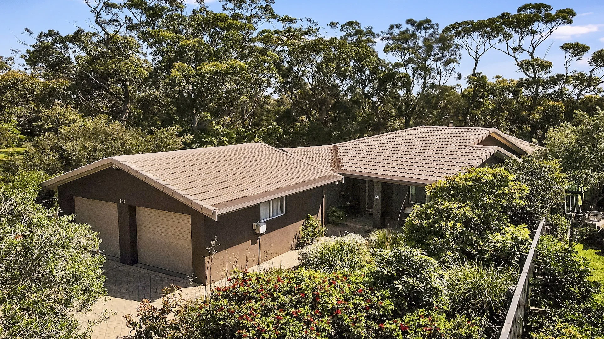 Reflections On the Creek house aerial shot of garage and house walls and a tiled roof surrounded by green trees and shrubs and a lush garden.