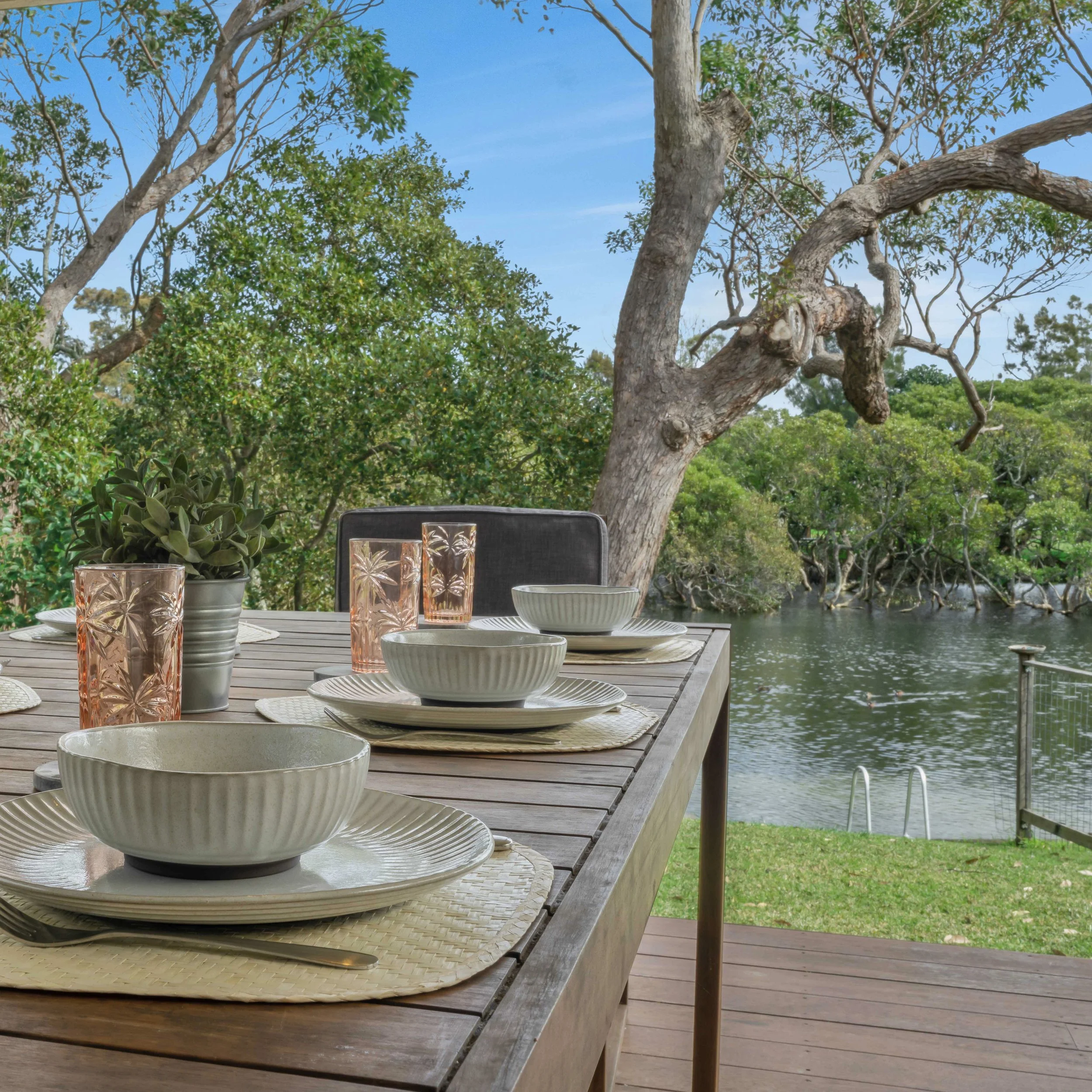 Outdoor dining table with white plates, pink-tinted glasses, and small potted plants, overlooking a tree-lined Currarong Creek under a clear blue sky.