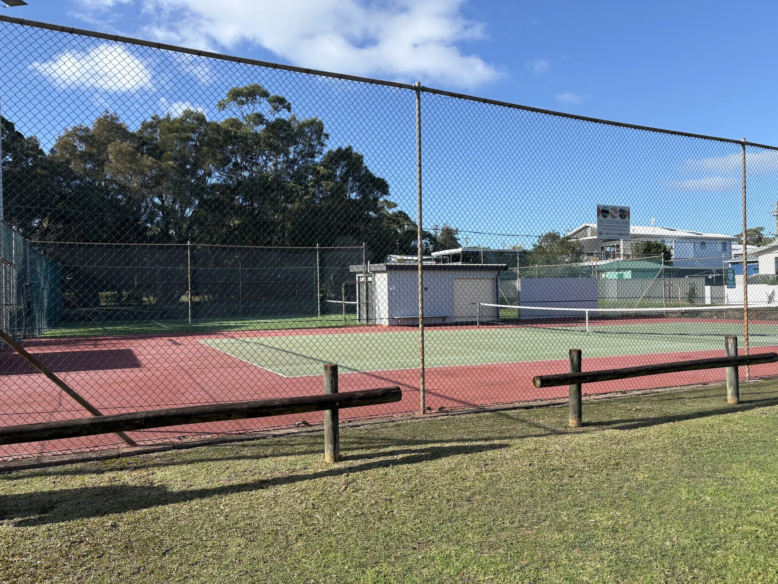 A tennis court surrounded by a chain-link fence with trees and buildings in the background under a partly cloudy sky.