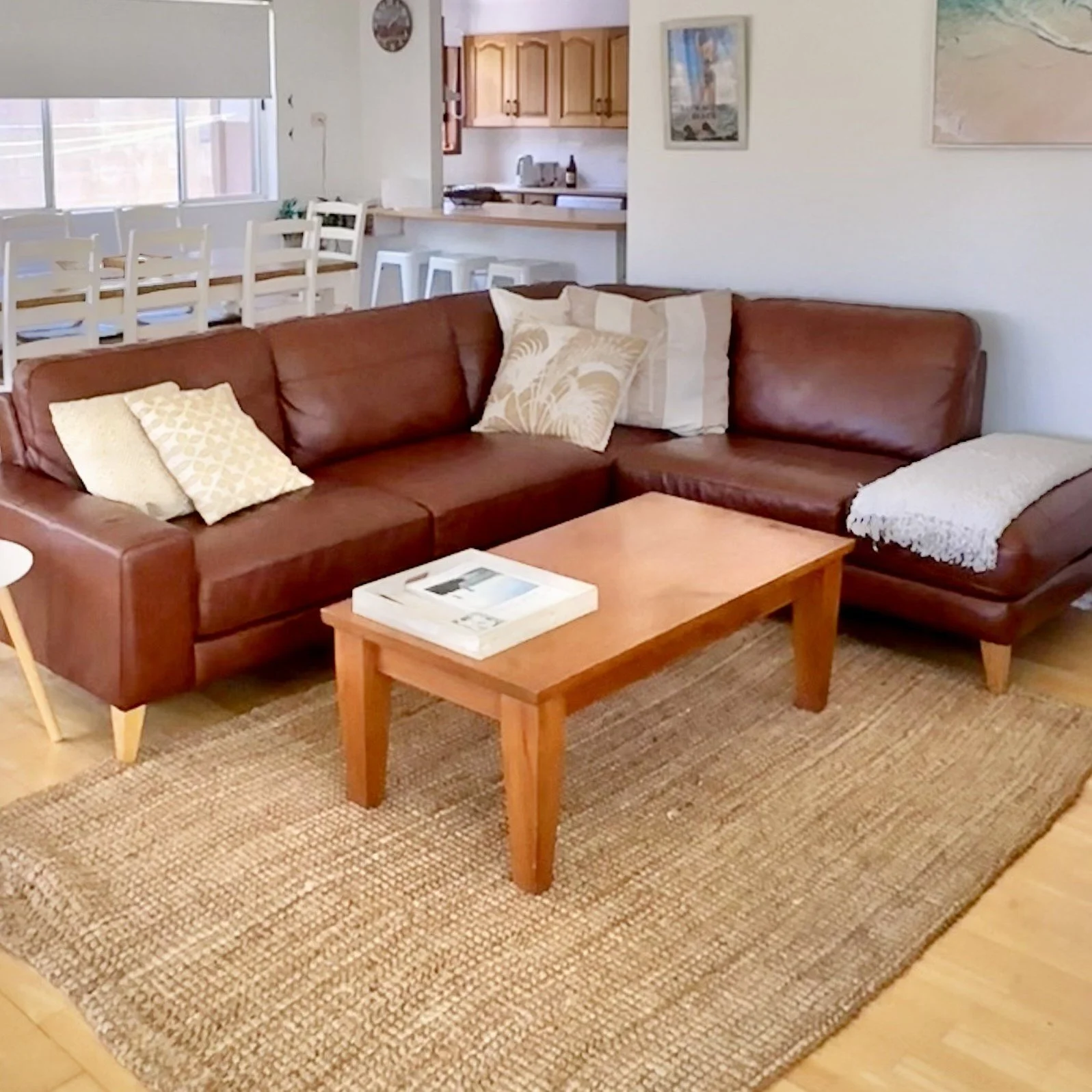 Living room with a brown leather L-shaped sofa, throw pillows, a wooden coffee table with a book, a beige rug, and a view into the kitchen with wooden cabinets, a breakfast bar with white stools, and pictures on the wall.