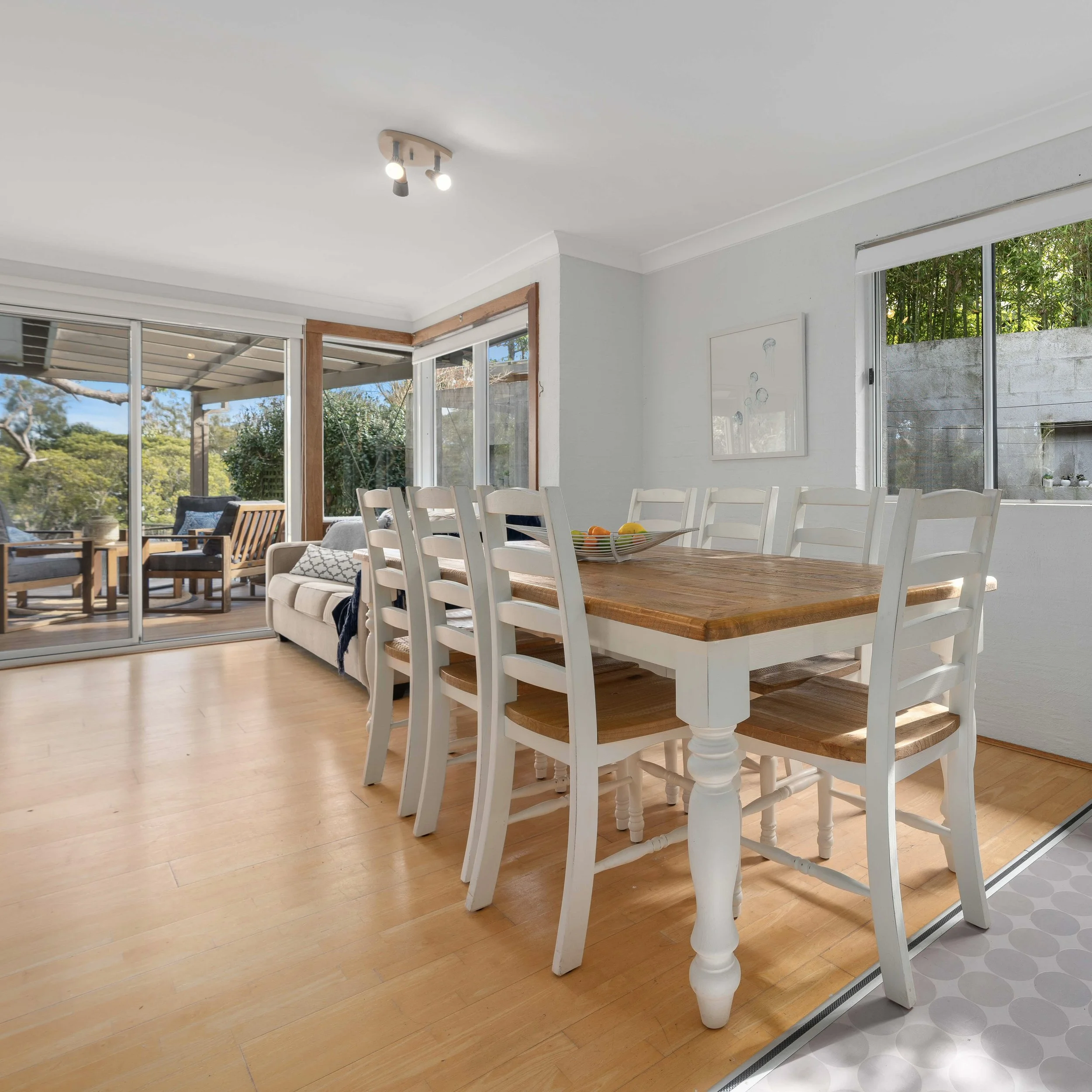 Living room with large windows, white walls, wooden dining table with white chairs, beige sofa, and outdoor greenery visible through the windows.