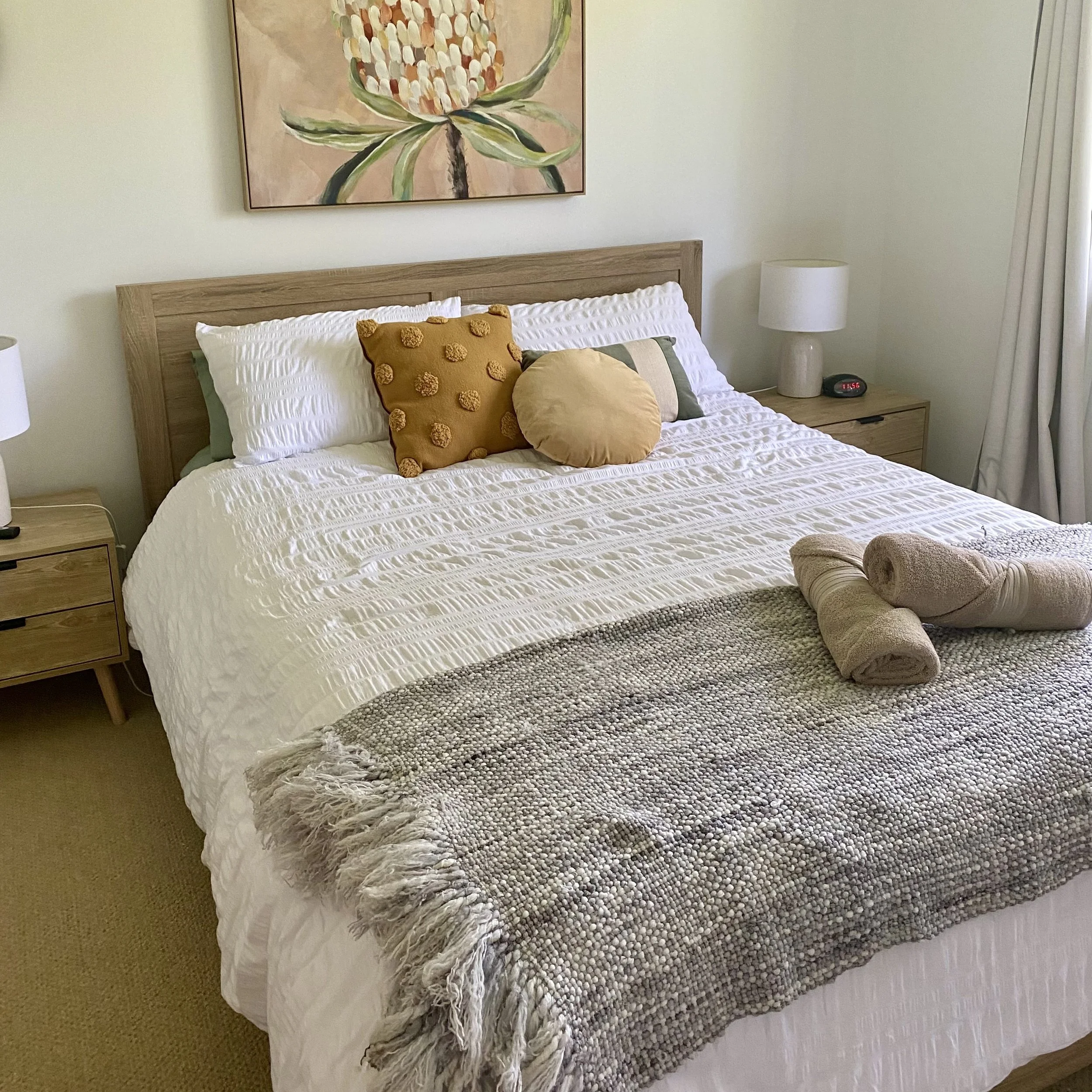 A neatly made bed with white textured bedding, accented by a mustard-colored pillow with pom-poms and a round beige pillow. The bed has a natural wood headboard and is flanked by matching wooden nightstands with white lamps. A large, abstract floral painting hangs above the bed, and a shaggy, textured throw blanket in gray and cream hues is draped at the foot of the bed.