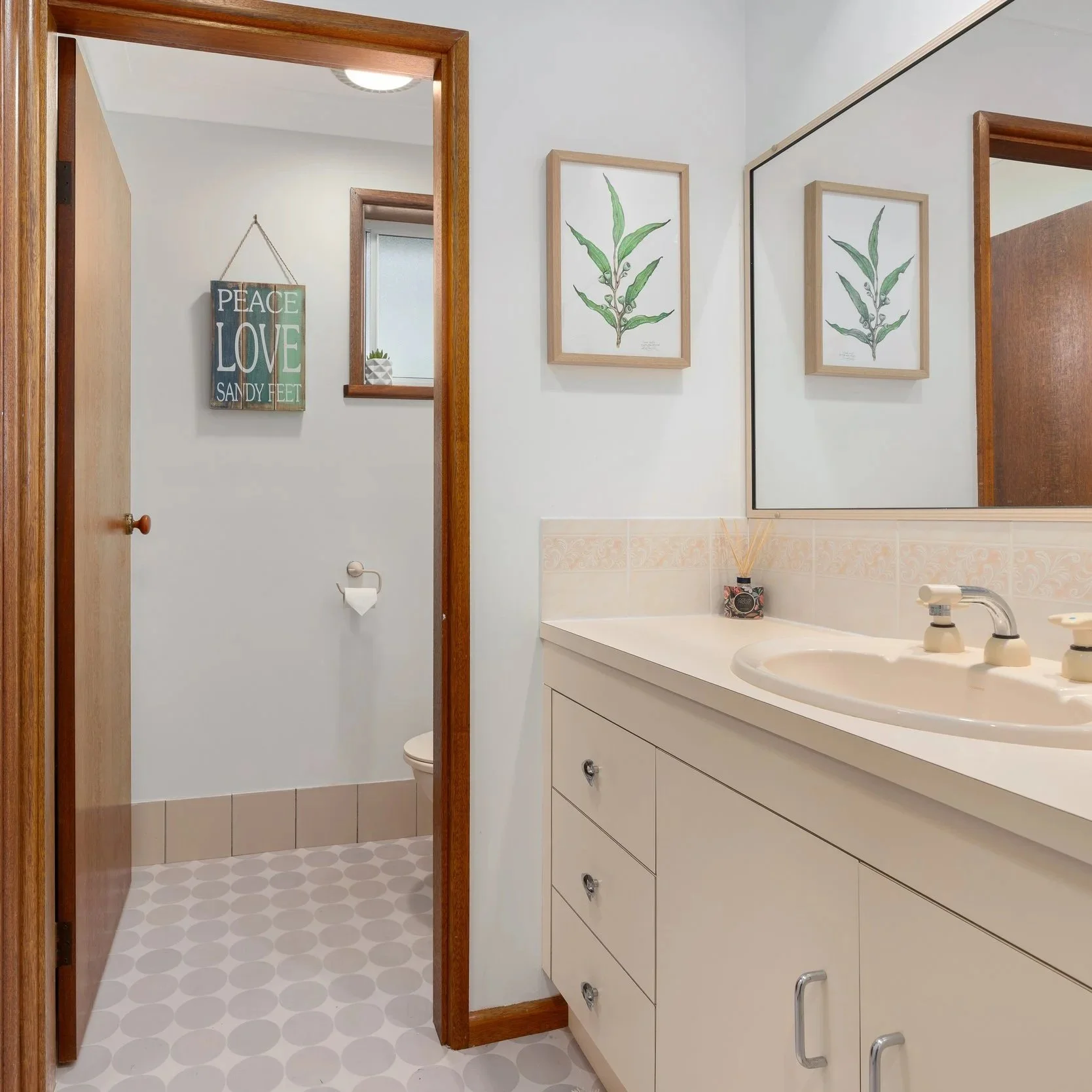 A bathroom with a white vanity sink, framed botanical artwork on the wall, and a small window with a decorative plant. The bathroom has a large mirror, a soap dispenser, and a framed sign that says 'Peace, Love, Sandy Feet' hanging in the toilet area.
