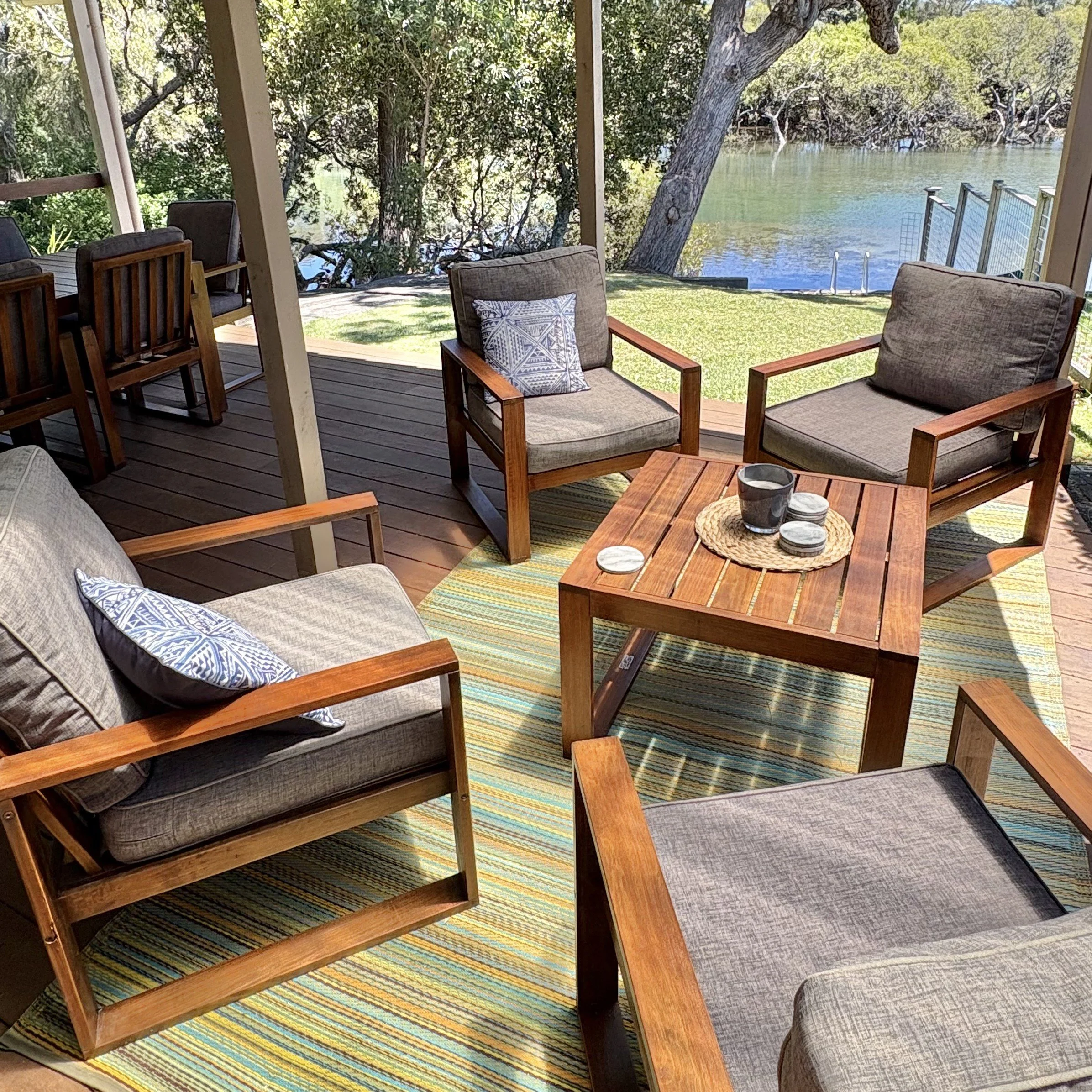 Indoor patio with outdoor furniture, including chairs and a sofa, overlooking a yard with green grass, trees, and a body of water with a ladder leading into it.