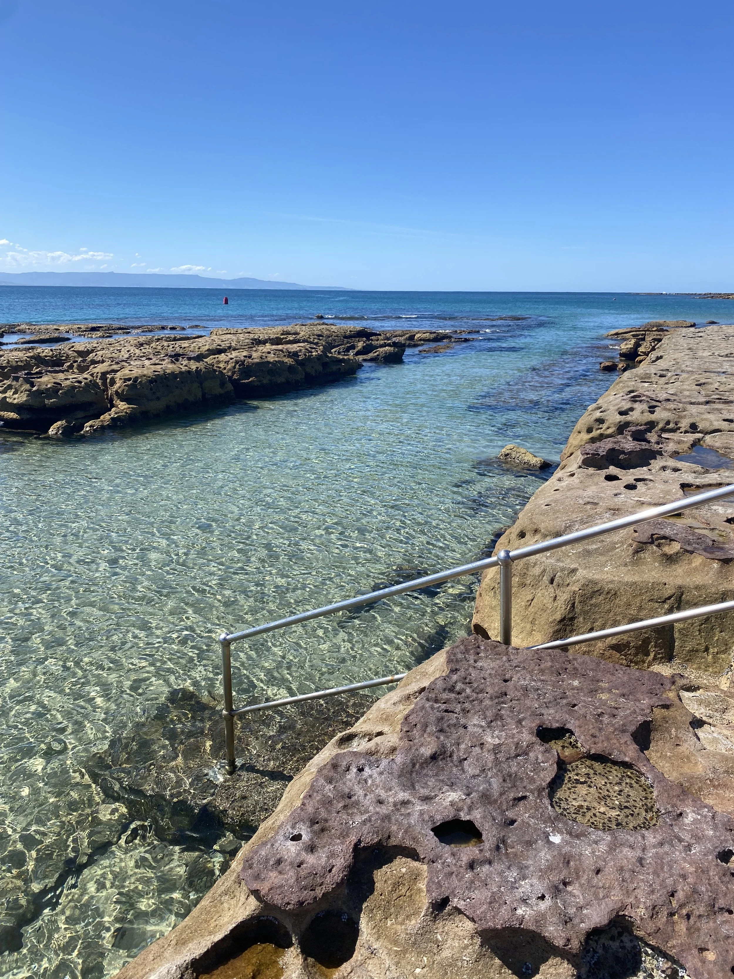 Clear shallow water with rock formations and a metal railing along a rocky shoreline, with the open sea and distant landmass under a bright blue sky.