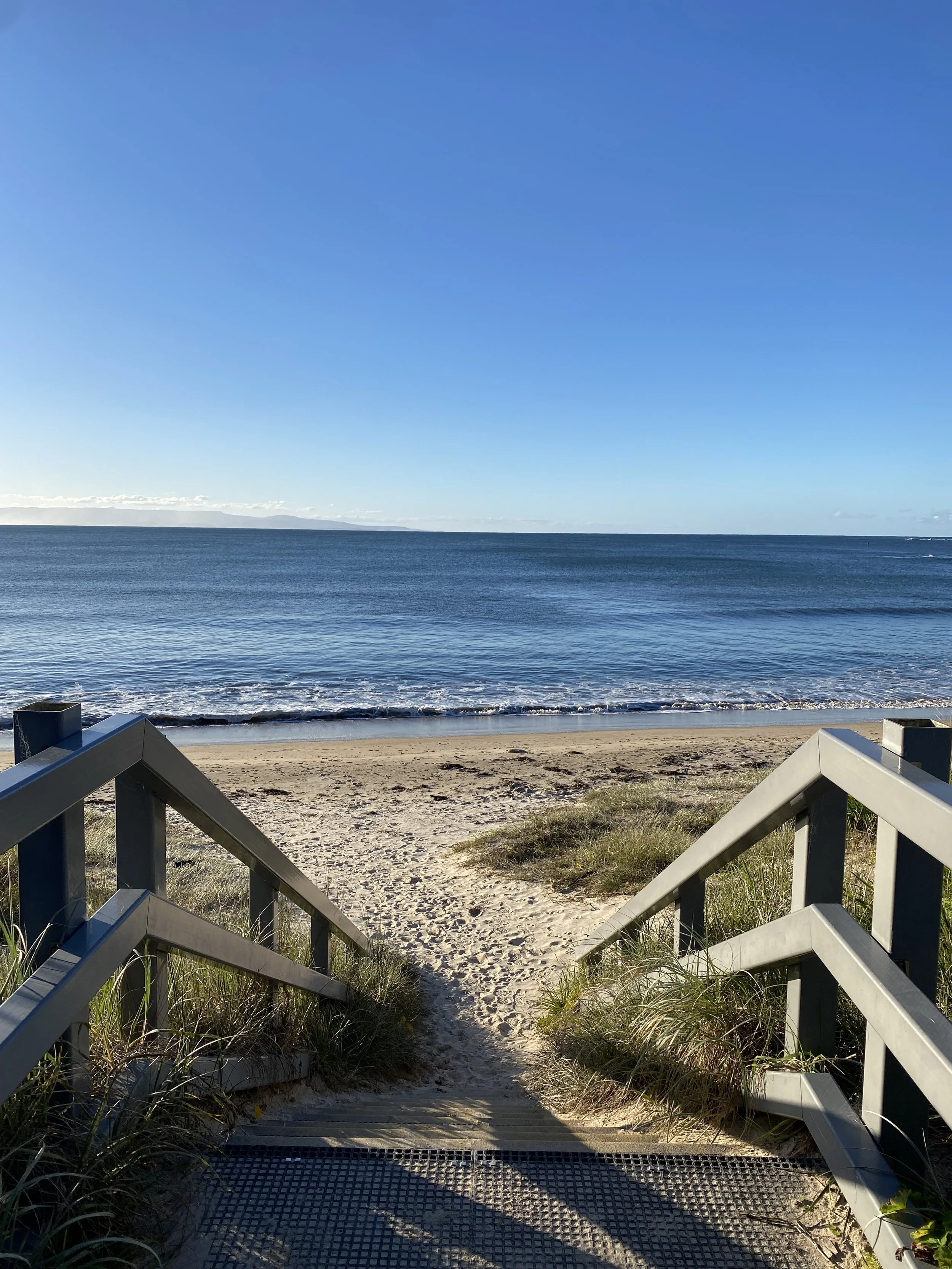 A staircase leading down to a sandy beach with the ocean and a clear blue sky in the background.