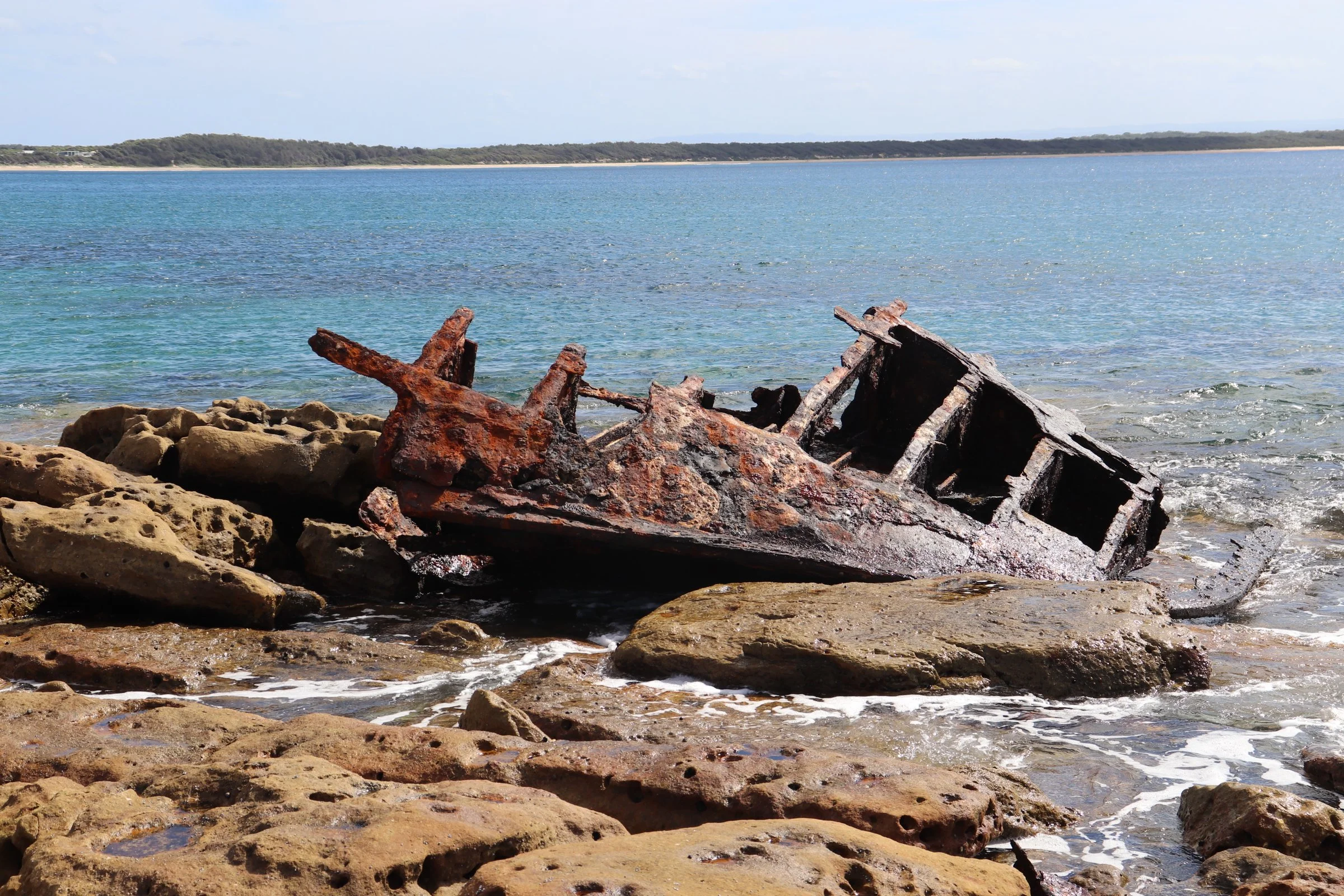 View of a rocky beach with rusty shipwreck debris in the foreground, calm blue water, and land with trees in the distance under a partly cloudy sky.