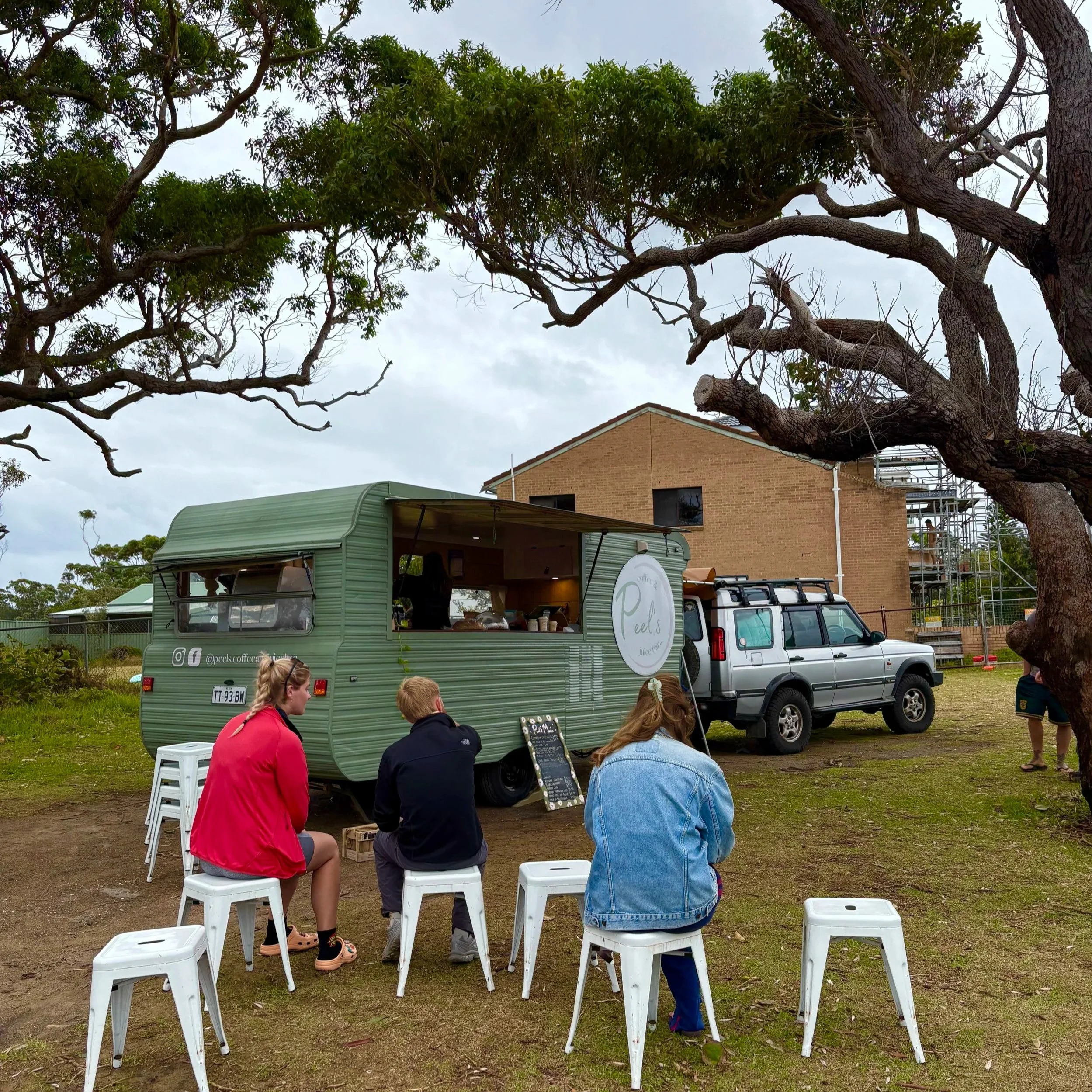 People sitting on white stools at Peel's Coffee and Juice bar in a vintage green caravan in a park in Currarong.