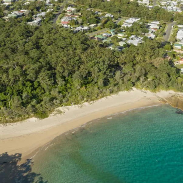 Aerial view of Abrahams Bosom Beach with white sand shoreline, turquoise water, and a residential neighborhood Currarong beyond.