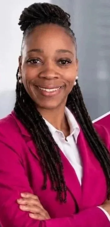 A woman with long braided hair, wearing a white collared shirt and a pink blazer, smiling with arms crossed against a neutral background.