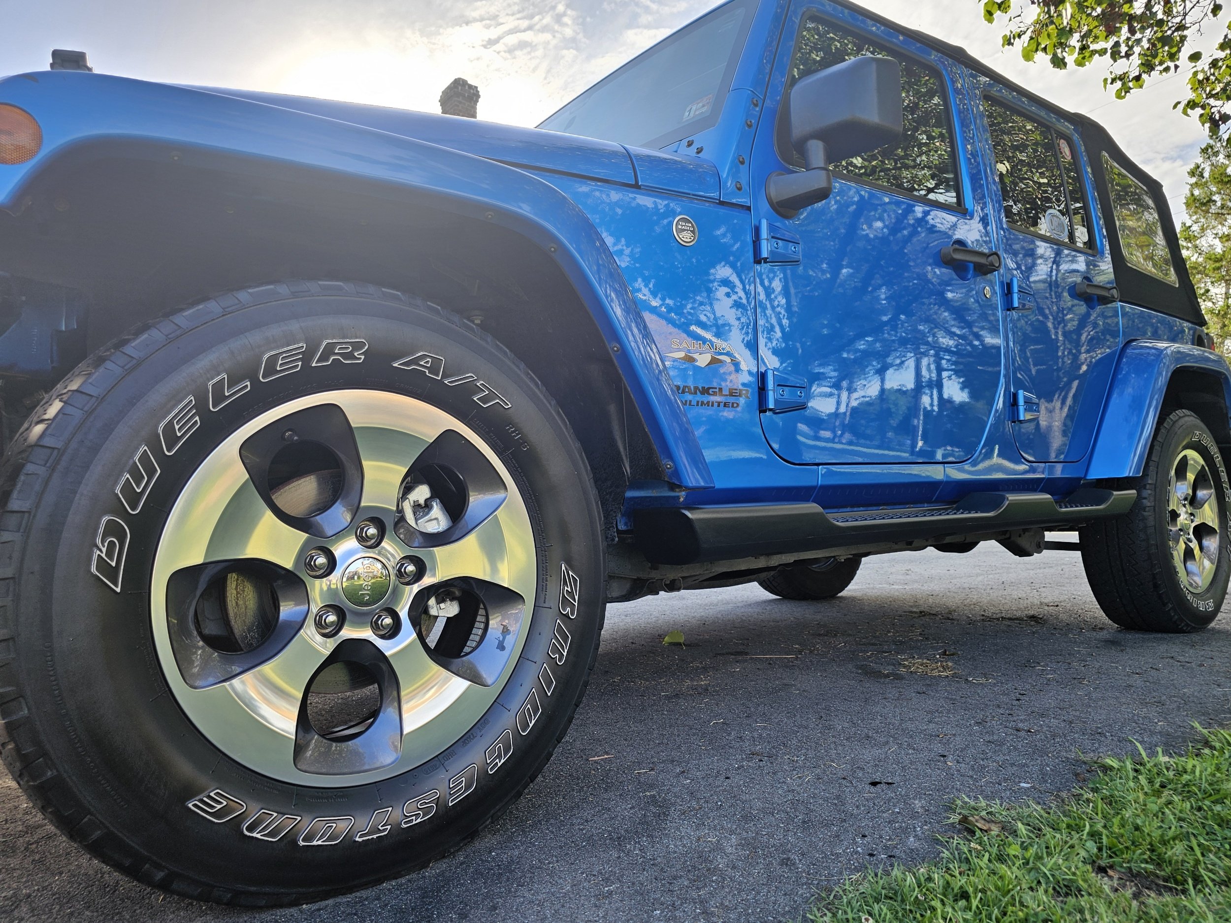 A blue Jeep Saharan Unlimited parked on pavement near grass area, with trees and cloudy sky in background.