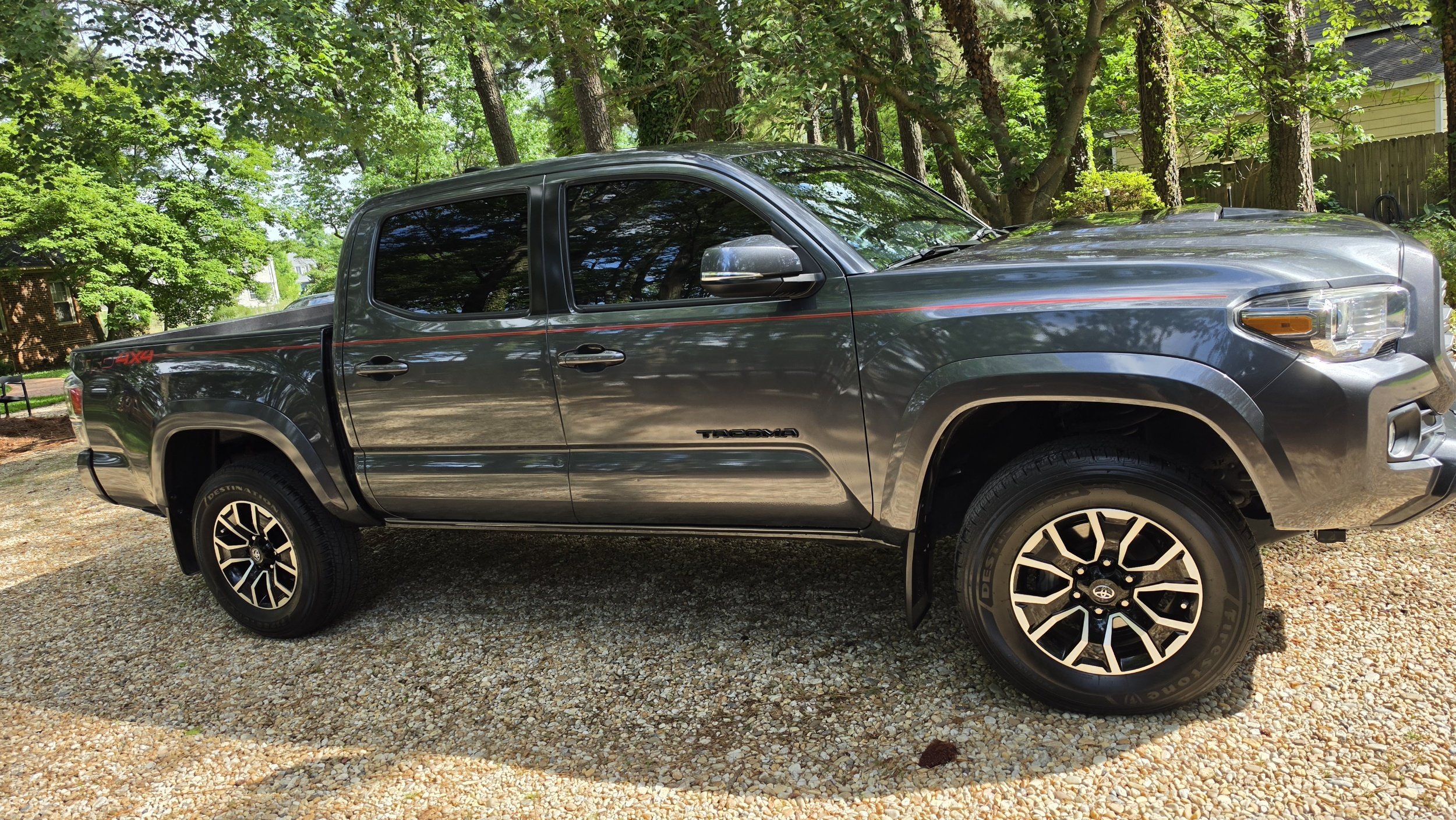 A gray Toyota Tacoma pickup truck parked on a gravel driveway, surrounded by green trees and houses in the background.