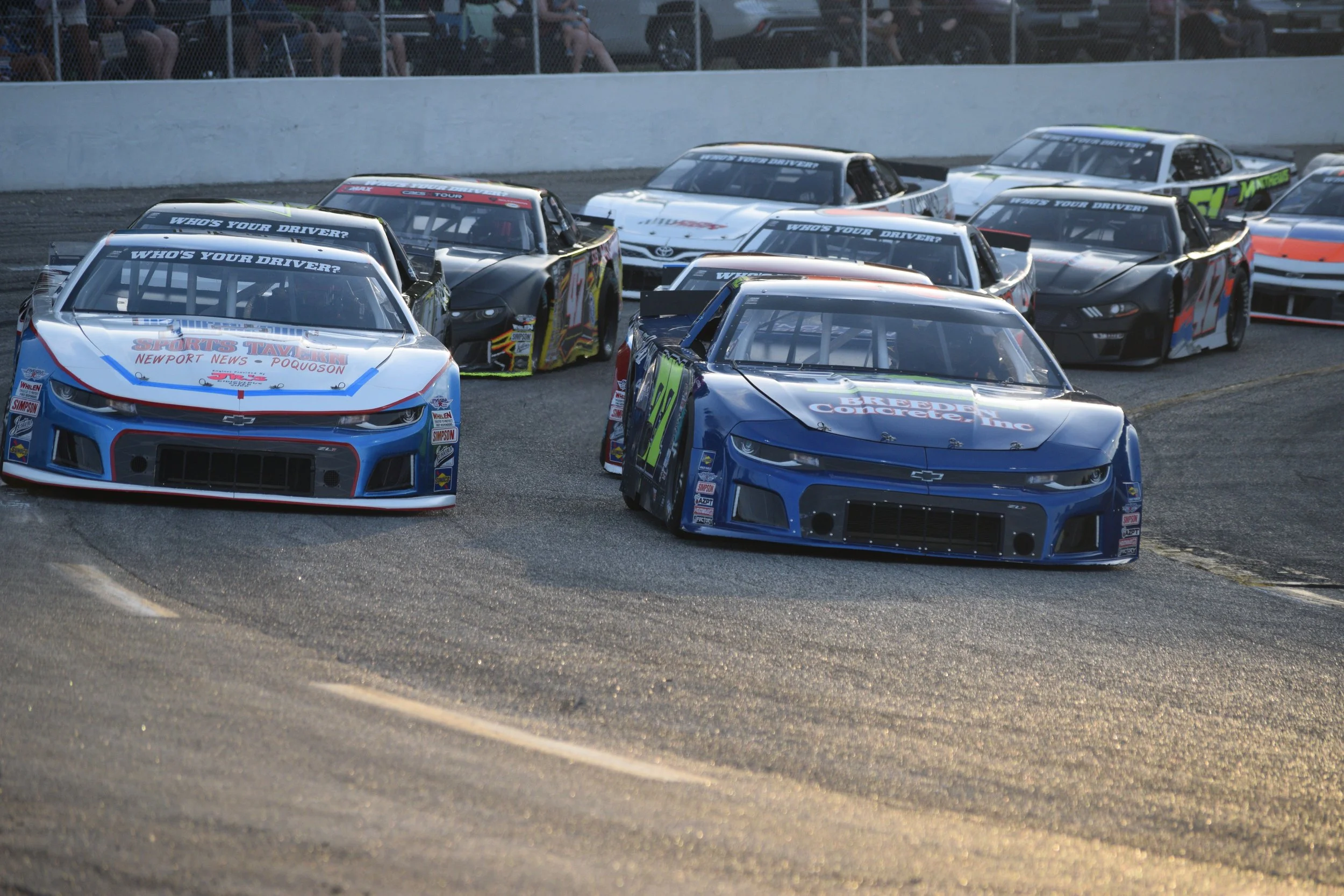 Multiple race cars on a racetrack, with spectators watching from the stands behind a fence.