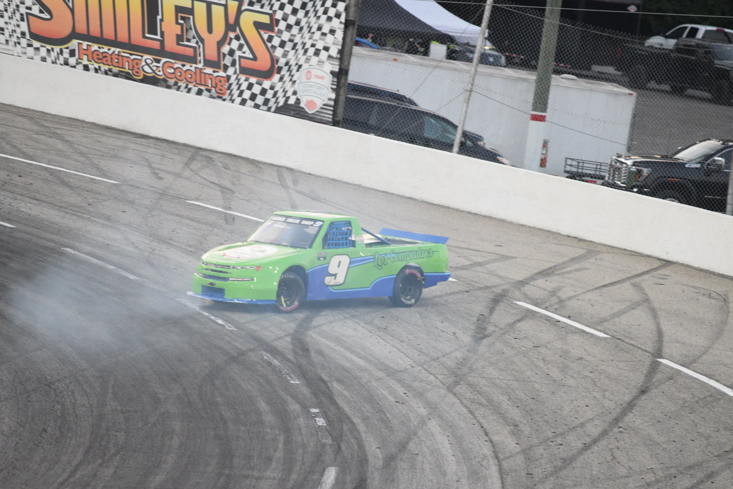 A green race car with the number 9 on its side speeding on a race track, with tire marks visible on the asphalt and a safety fence in the background.