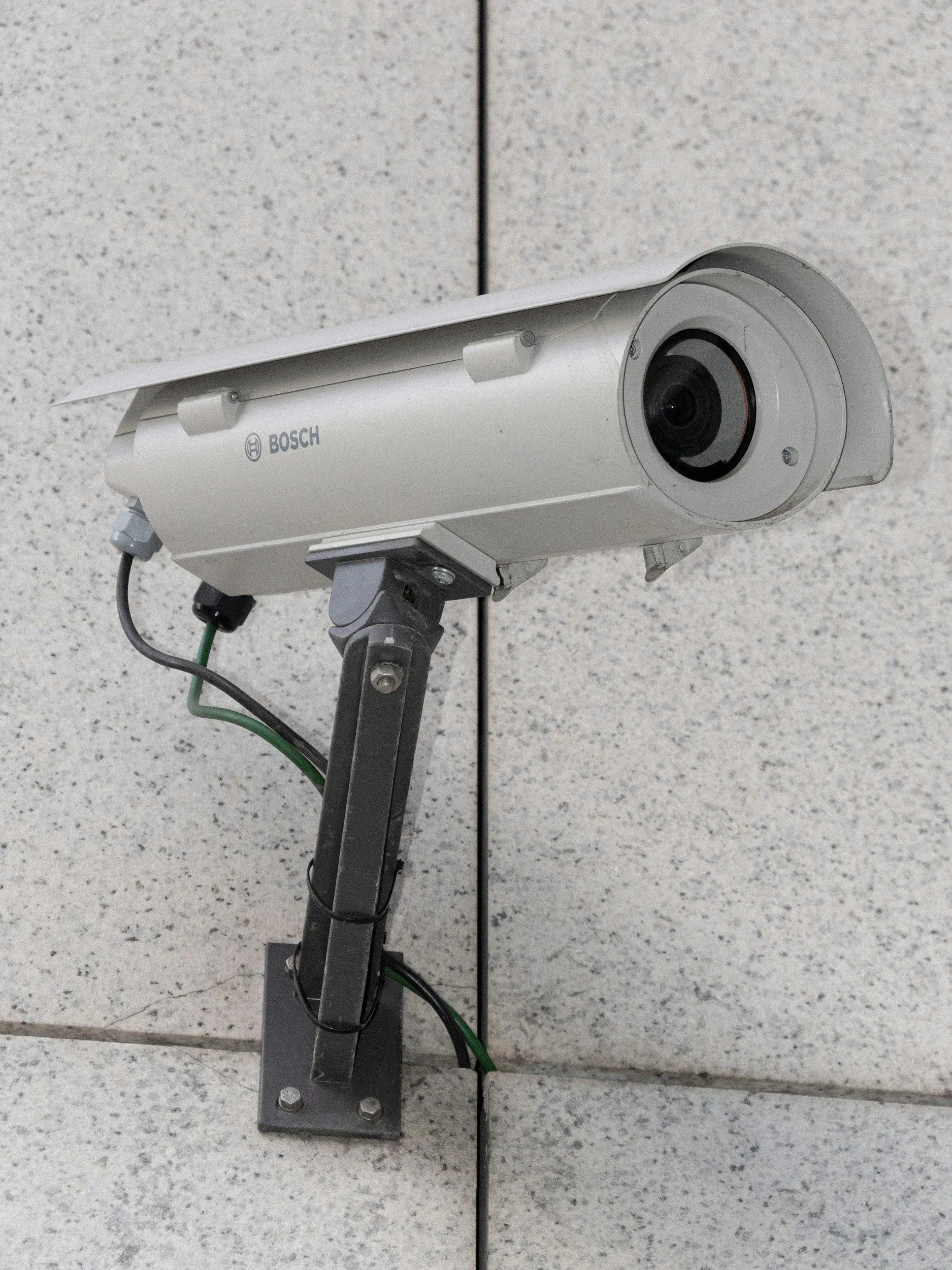A white security camera mounted on a metal stand on a speckled gray tiled wall.