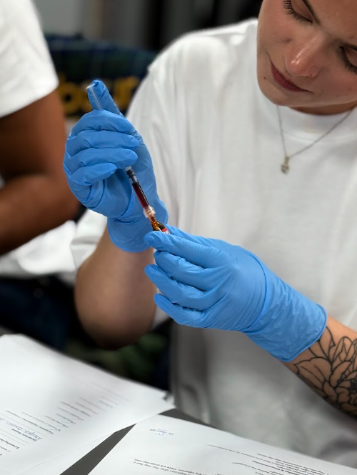 A person is preparing to give an injection to a model of a knee, with a woman sitting at a table observing.
