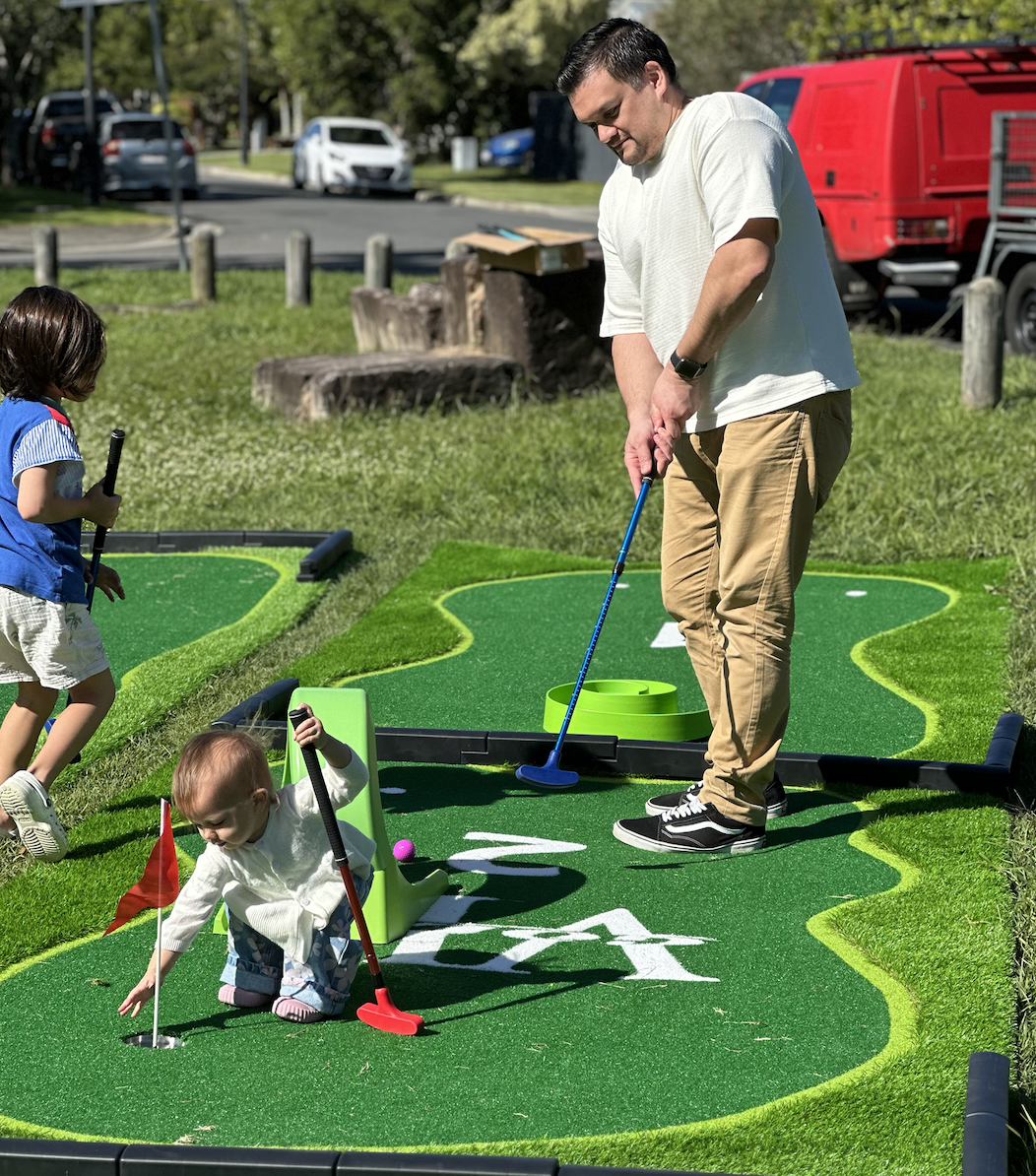 A man and two children playing miniature golf outdoors on a sunny day, with boats and cars in the background.