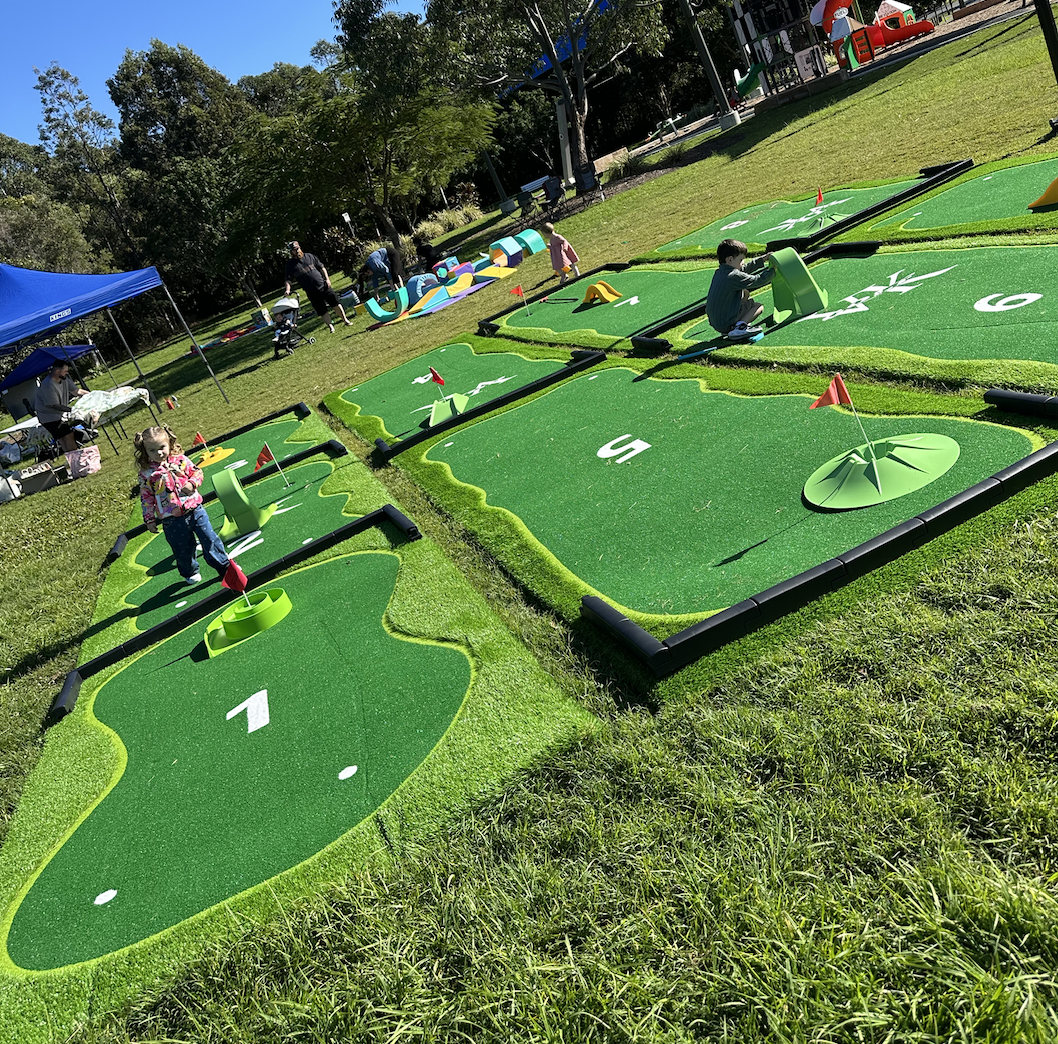 Miniature golf course with multiple holes, each with putting greens, flags, and obstacles. Children and adults are playing and walking around under a clear blue sky, with trees and a playground in the background.