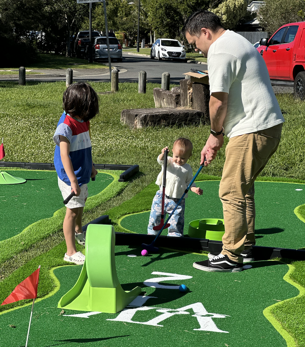 A man and two children playing mini golf outdoors on a sunny day. The man is helping a young child with a golf club, while another child watches nearby.