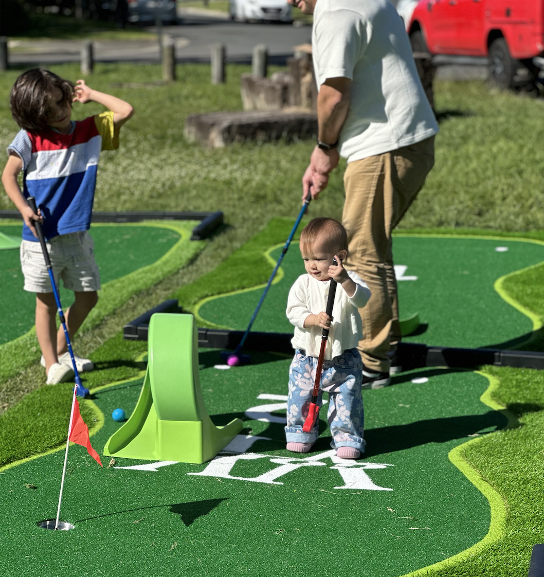 Children playing mini golf outdoors on a sunny day, with a man supervising, on a course with green artificial turf and decorative winding borders.