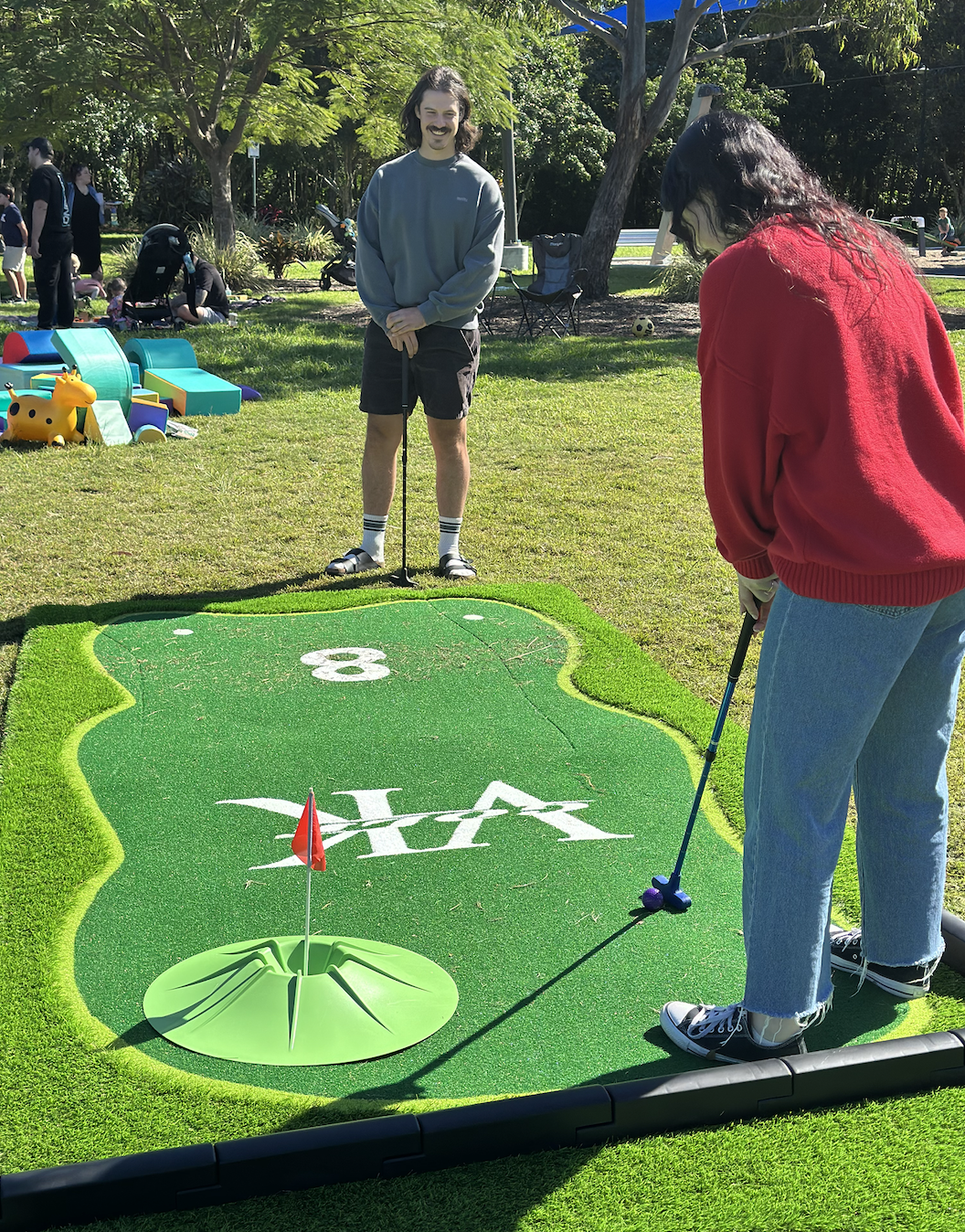 Two women playing miniature golf outdoors on a sunny day, with one about to take a shot and a man watching, while other people relax and play in the background.
