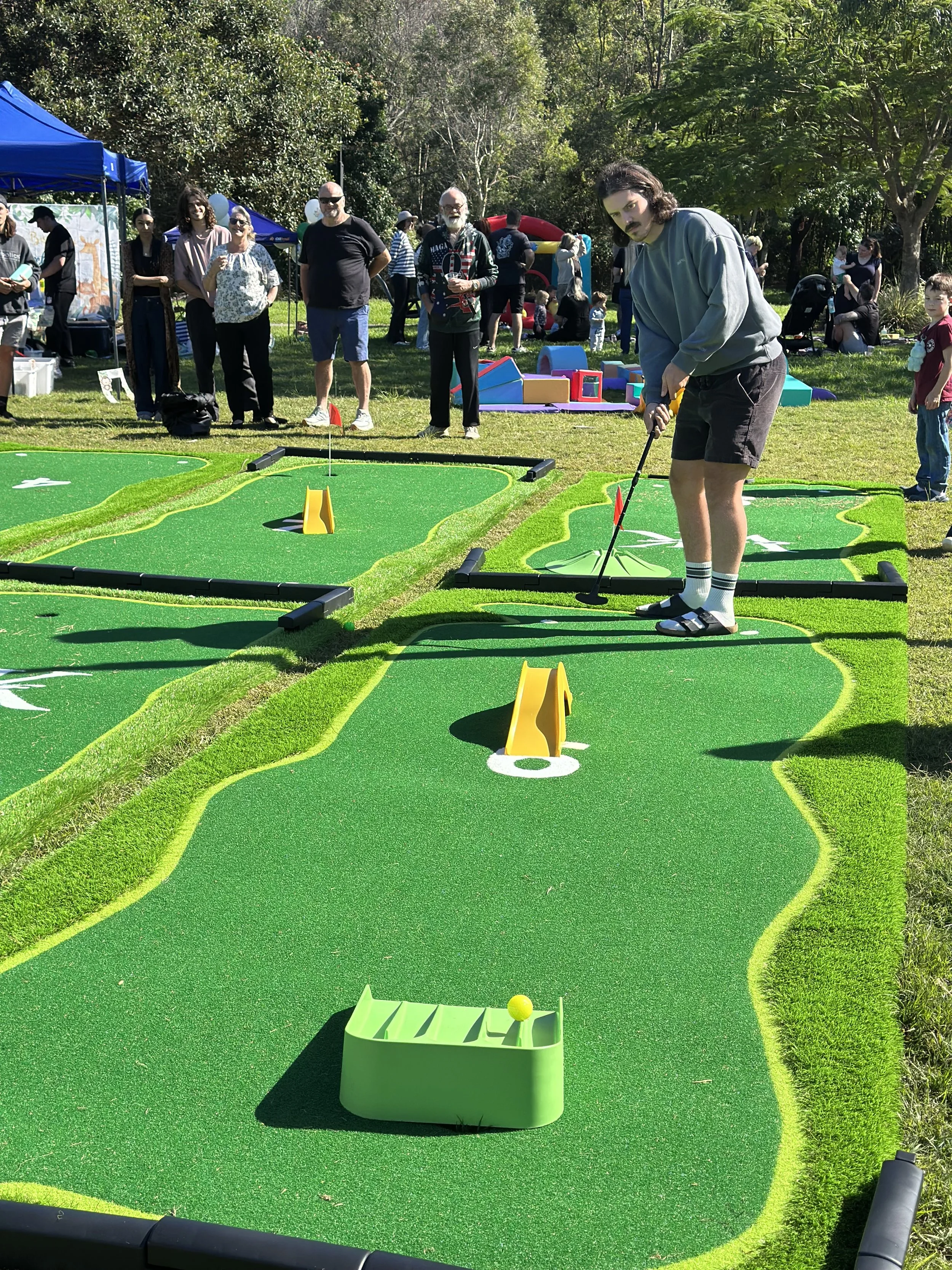 People gathered outdoors on a sunny day at a mini golf course. A young man is preparing to hit a golf ball on a green mat, while others stand and enjoy the scene. There are tents, children playing, and trees in the background.