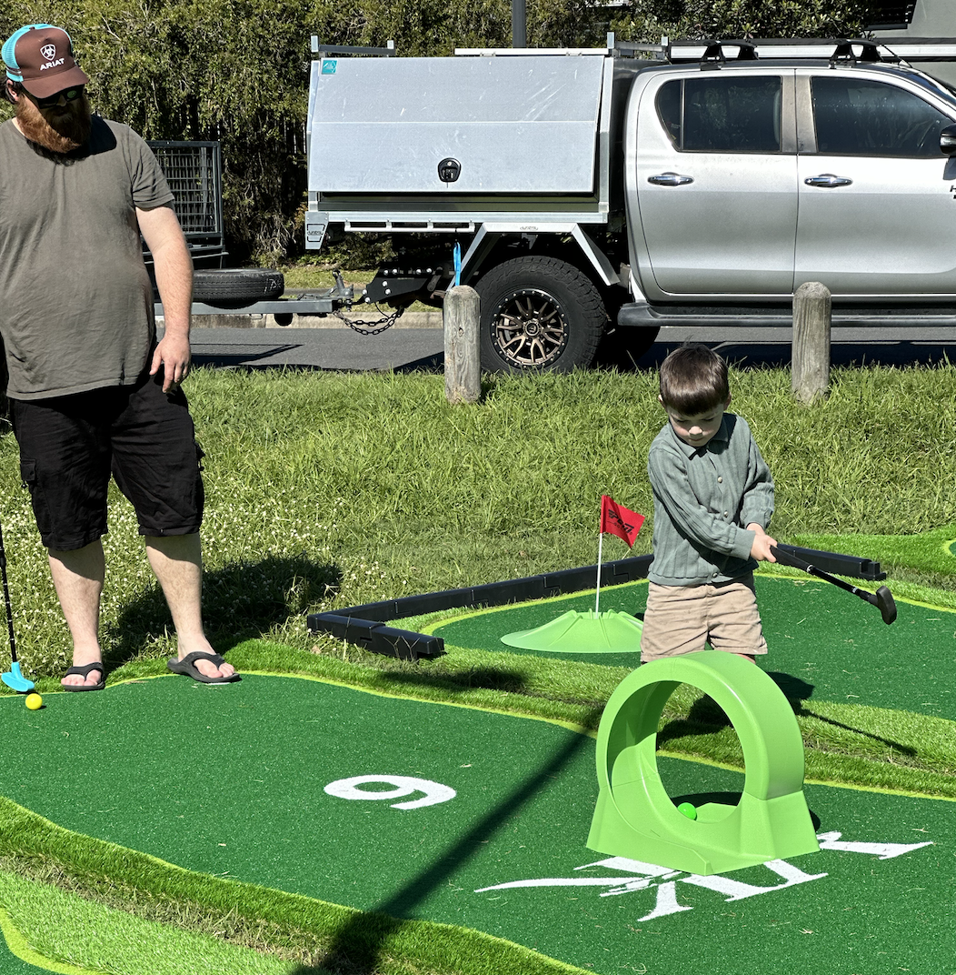 A man and a boy playing miniature golf on a green course with a small hill and a red flag. The man is wearing a gray T-shirt, black shorts, flip-flops, and a cap, and he is standing near the edge of the course. The boy is wearing a gray shirt and tan