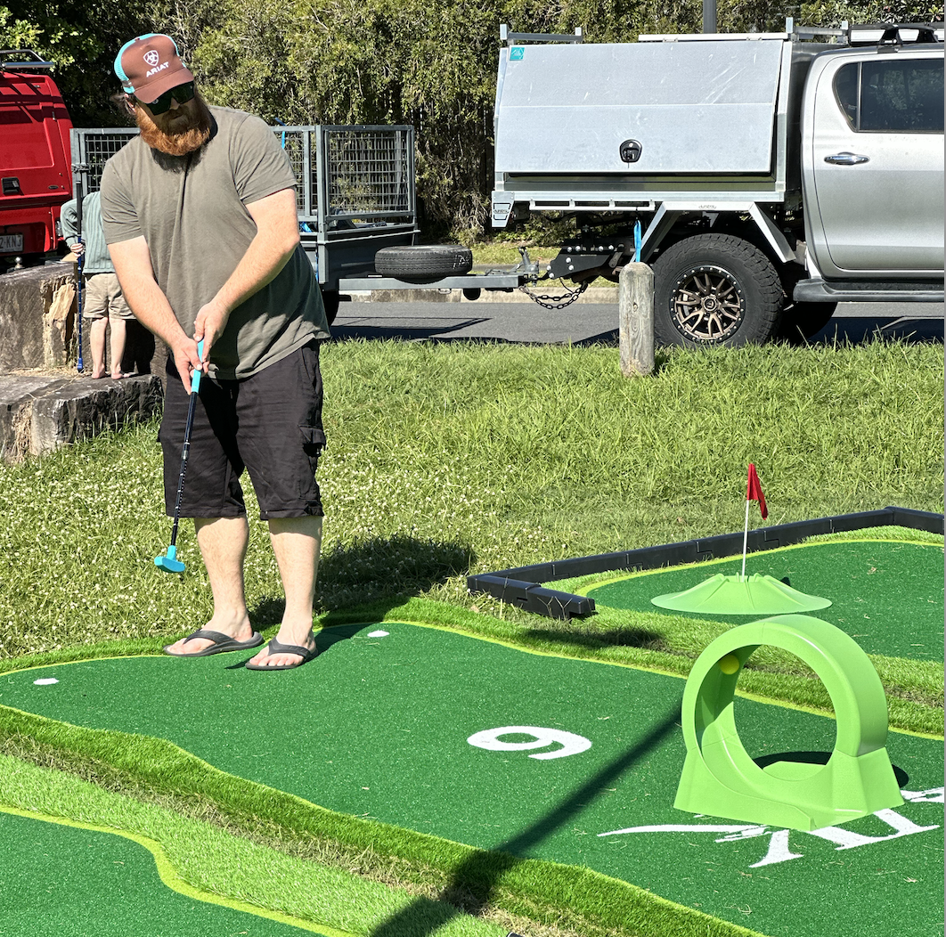 Man playing mini golf outdoors on a bright sunny day, standing on a green artificial turf putting area, holding a putter, with a golf hole marked with a flag in the background, near parked trucks and greenery.