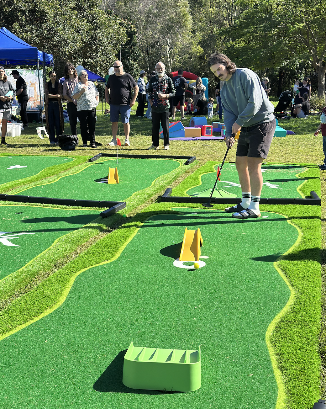 People playing mini golf outdoors with green turf and obstacles, surrounded by trees and other participants.