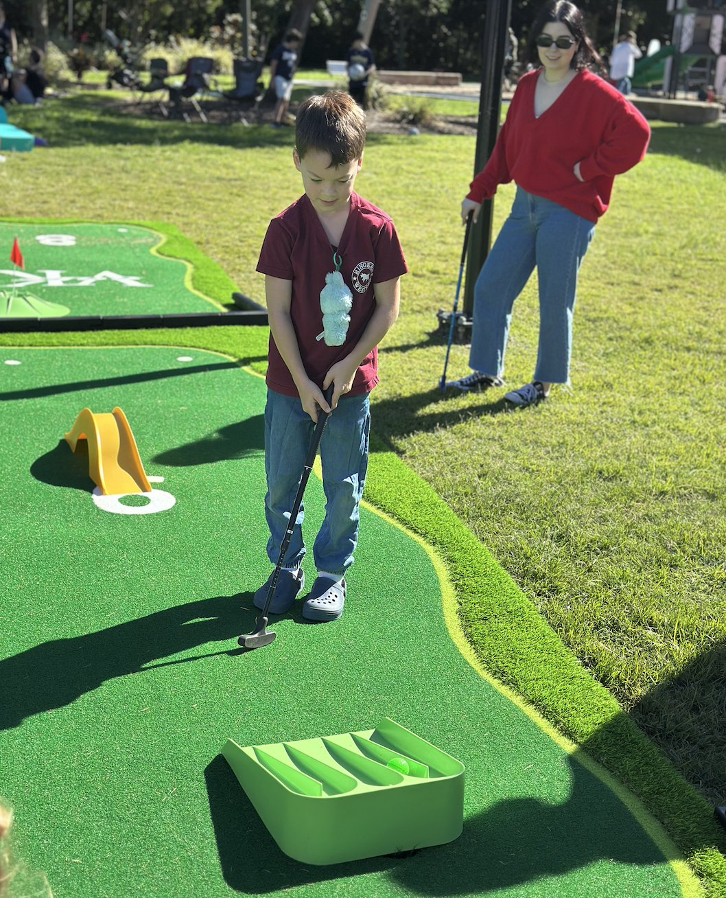 A boy and woman playing mini golf outdoors on a sunny day.