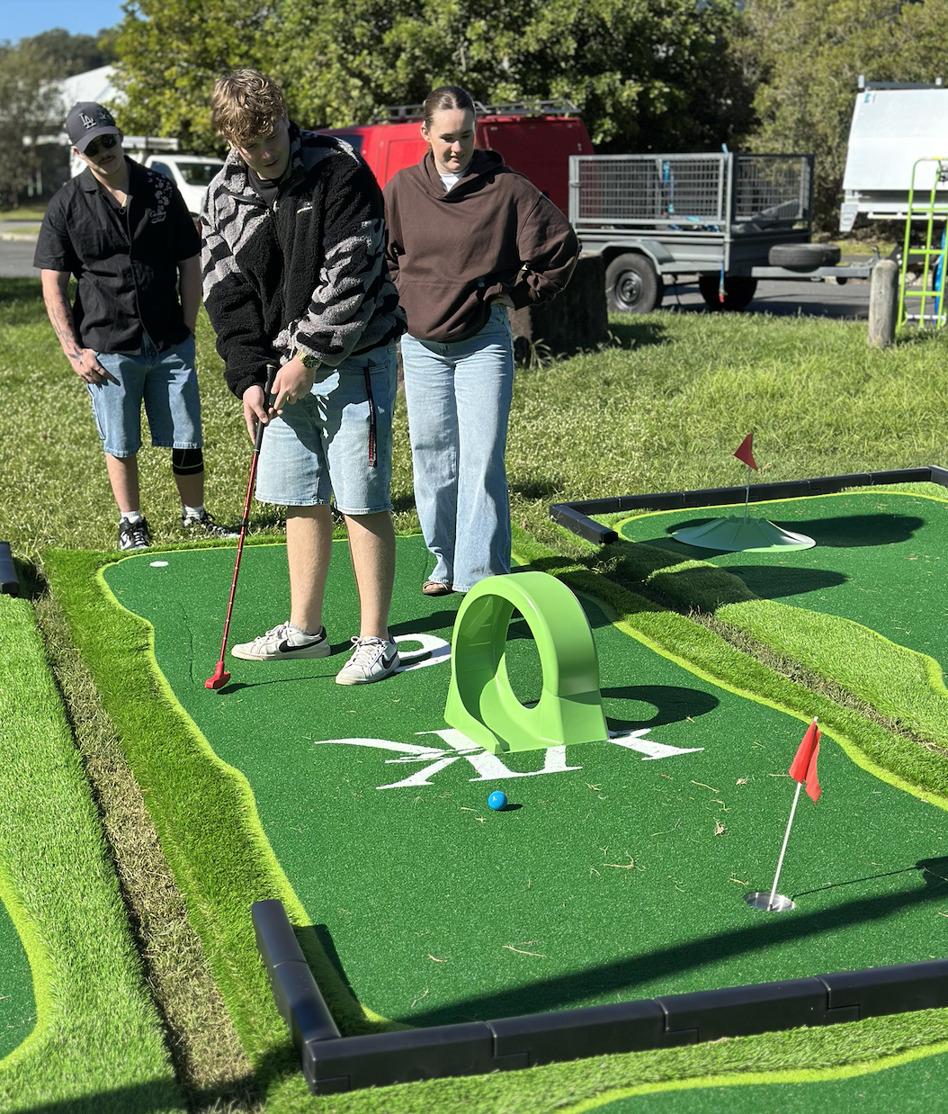 Three people playing miniature golf on a sunny outdoor course with green artificial turf, small flags, and a hole with a cover. One person is preparing to hit the ball while two others watch.