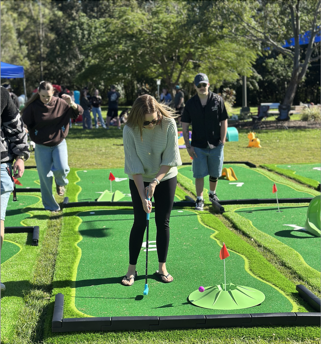 People playing miniature golf outdoors on a sunny day with green artificial turf, small hills, and flags on the course.