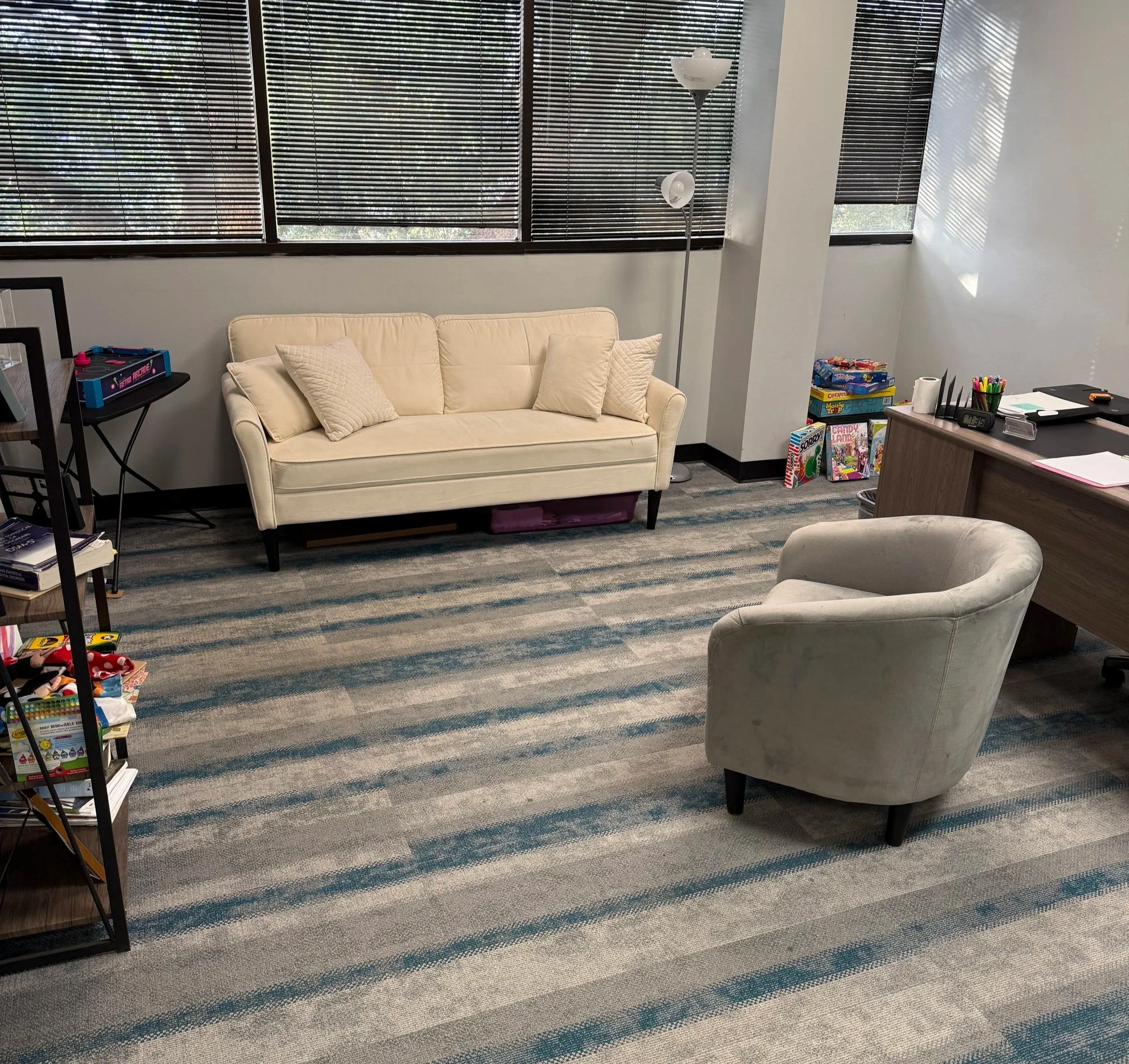 A cozy office space with a cream sofa, beige pillows, a standing floor lamp, and a large window with closed blinds letting in filtered sunlight. There are various board games and office supplies on a desk and shelves.