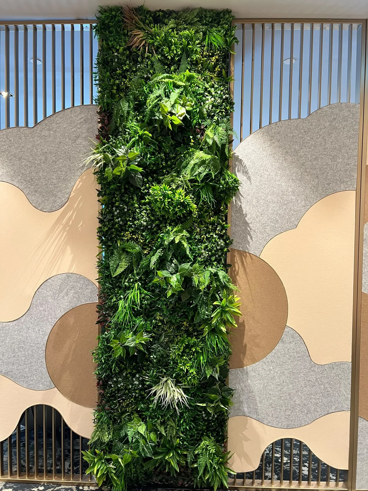 A vertical green plant wall with various types of lush, leafy plants, framed by a decorative wooden grid with colorful, abstract shapes on beige and gray background panels.