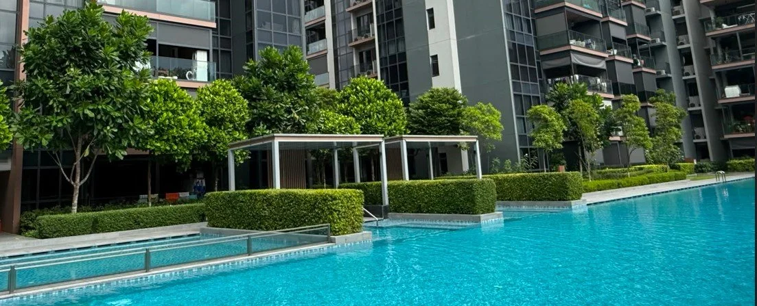 Modern residential apartment complex with a large swimming pool, surrounded by trimmed hedges and green trees.