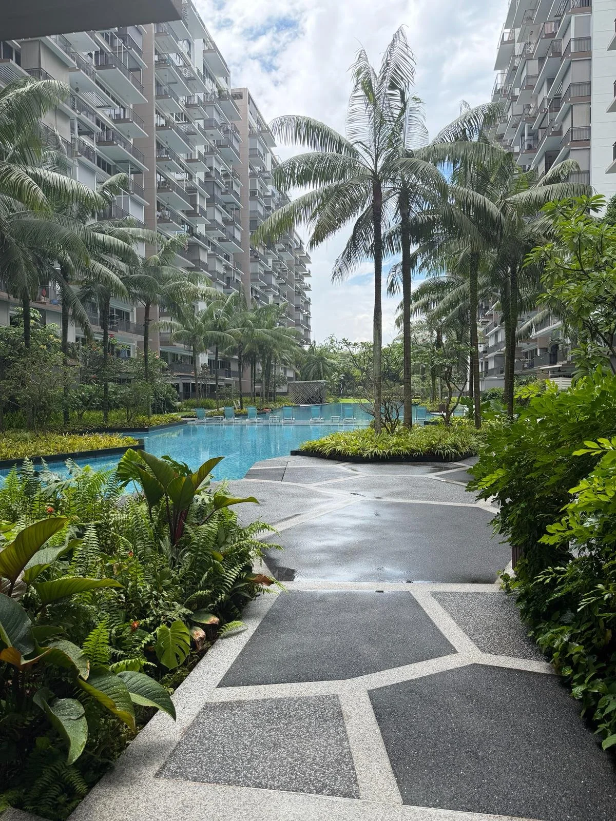 A luxurious residential complex with lush tropical plants, palm trees, a swimming pool, and high-rise apartment buildings in the background under a partly cloudy sky.