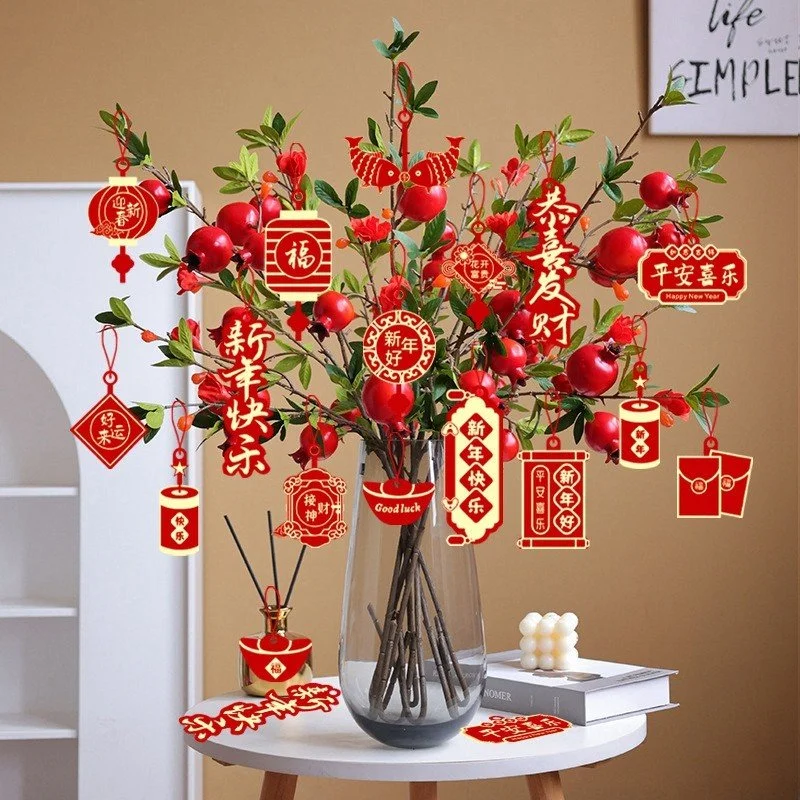 Decorative Chinese New Year arrangement in a glass vase with red and gold paper ornaments hanging from tree branches, featuring traditional symbols and greetings, on a white round table with a cube-shaped candle, books, and a red cut-out message